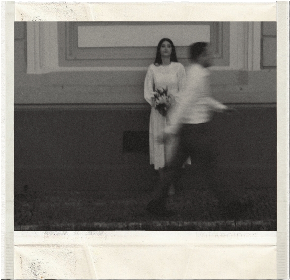 A woman standing against a wall holding flowers, with a man walking past quickly in front of her.