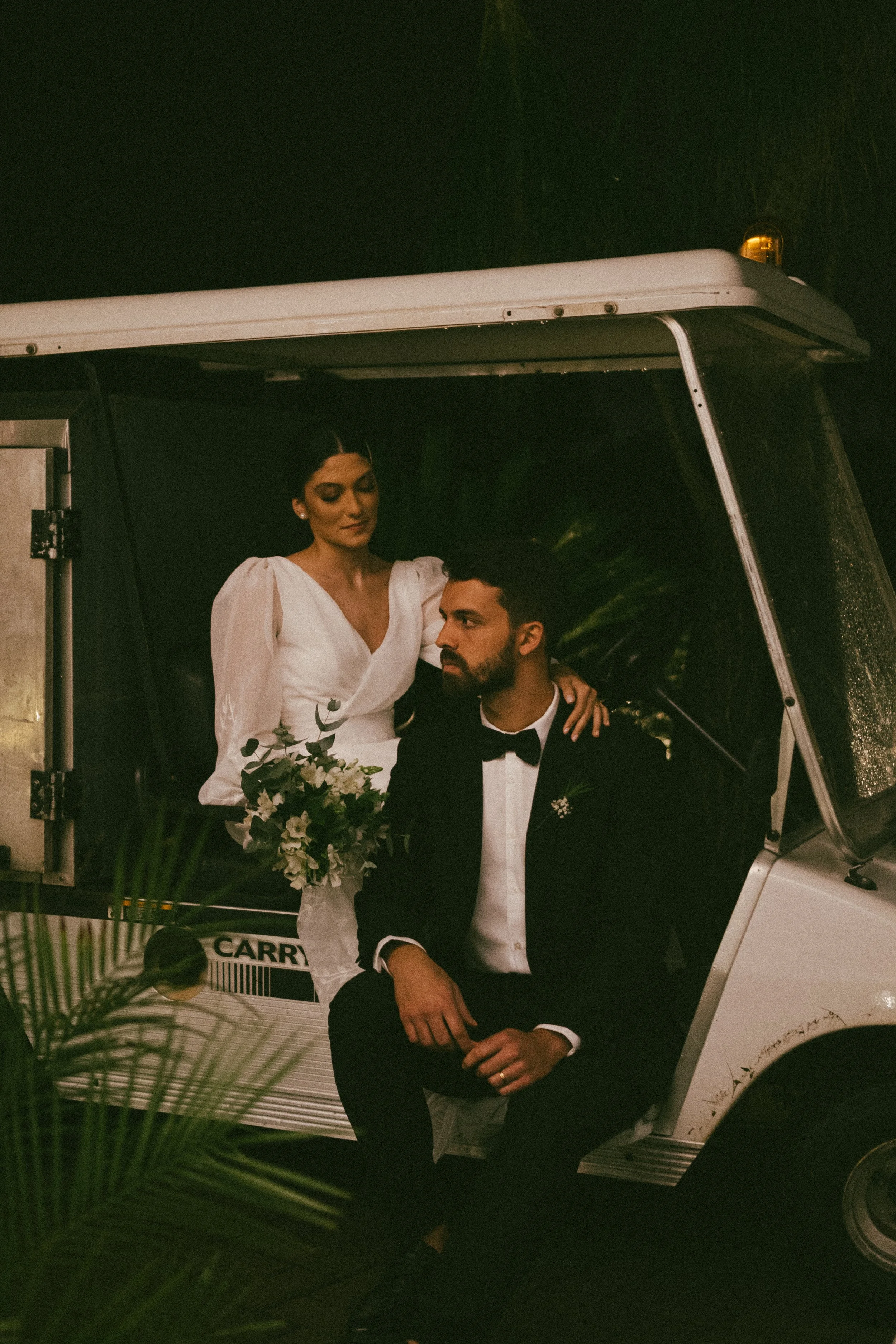 A bride and groom sitting on an ice cream cart, with the bride holding a bouquet of white flowers, at night surrounded by greenery.