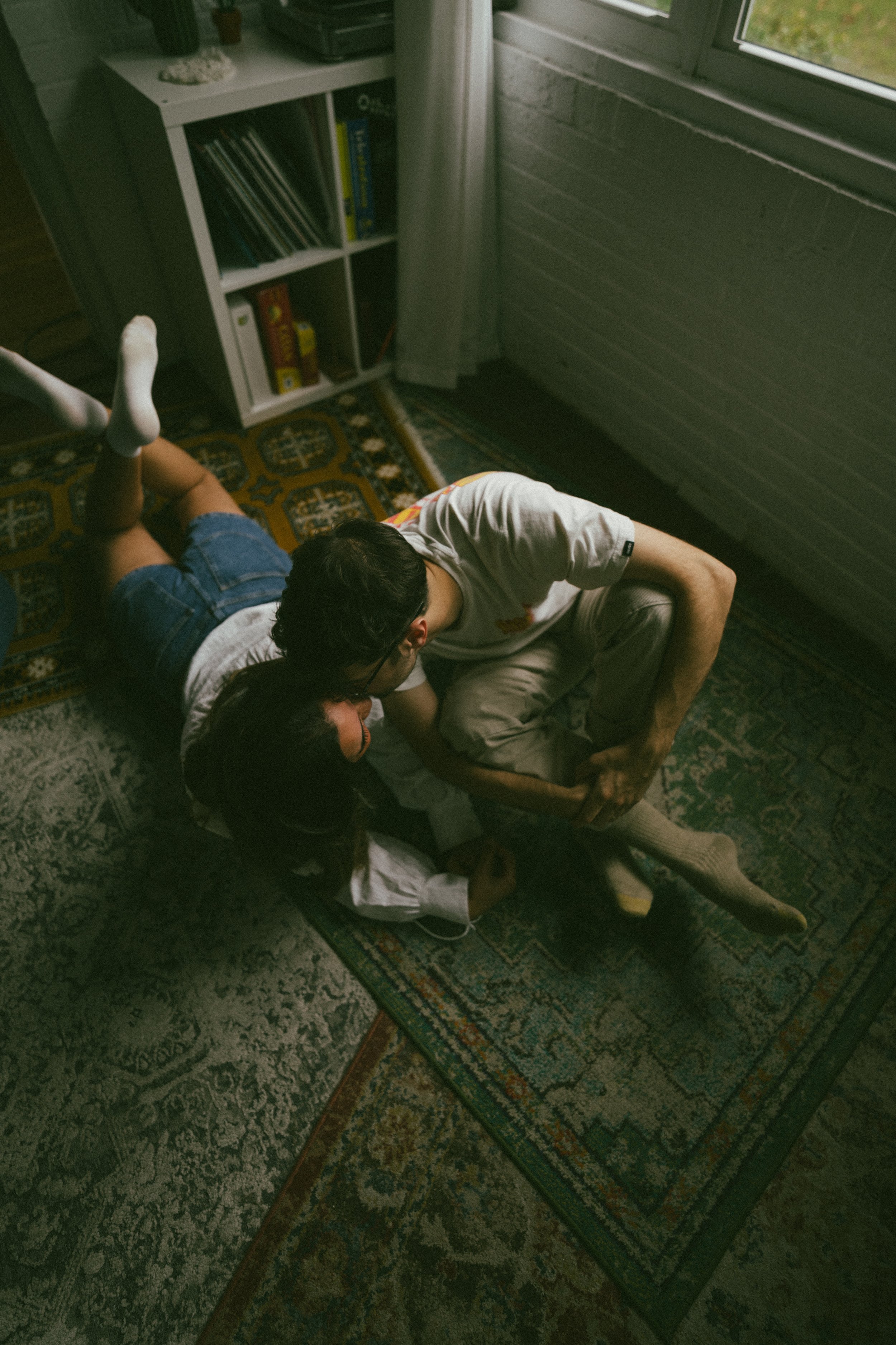 Two children and a man are on the floor near a window, with bookshelves in the background. The children are lying on the carpet, and the man is sitting on a small chair. They appear to be playing or cuddling.