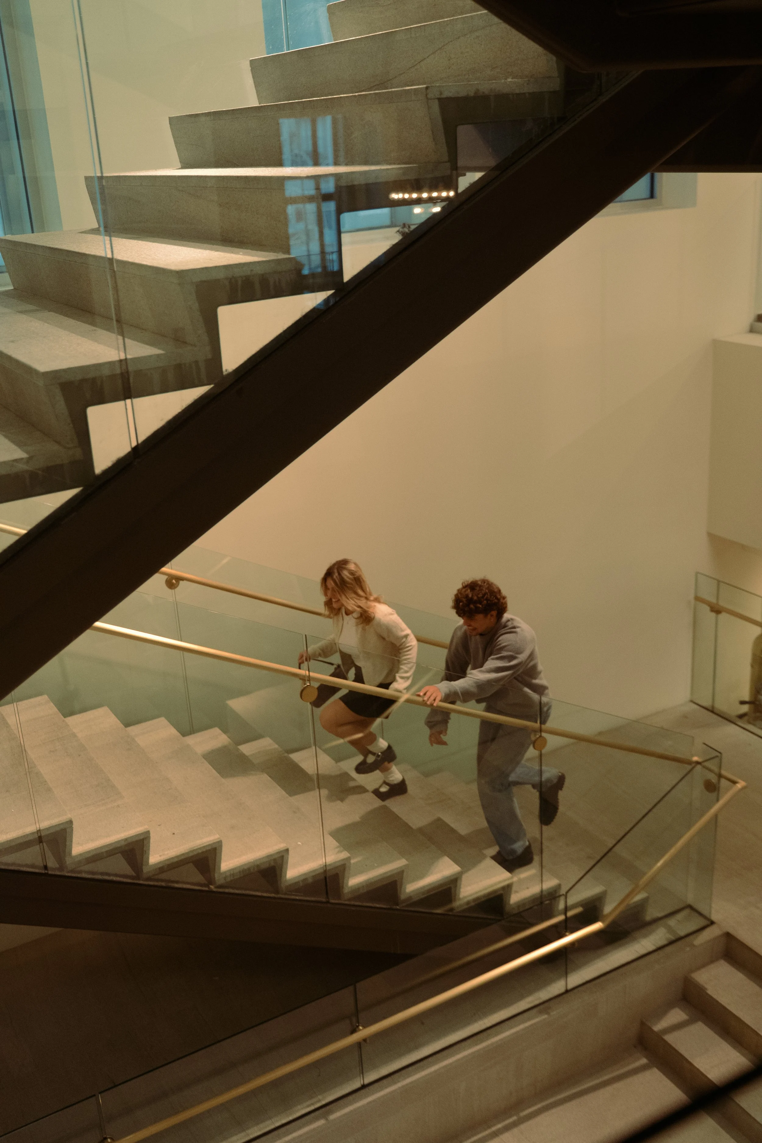 Two people, a woman and a man, are climbing up a stairwell with glass railings inside a modern building.