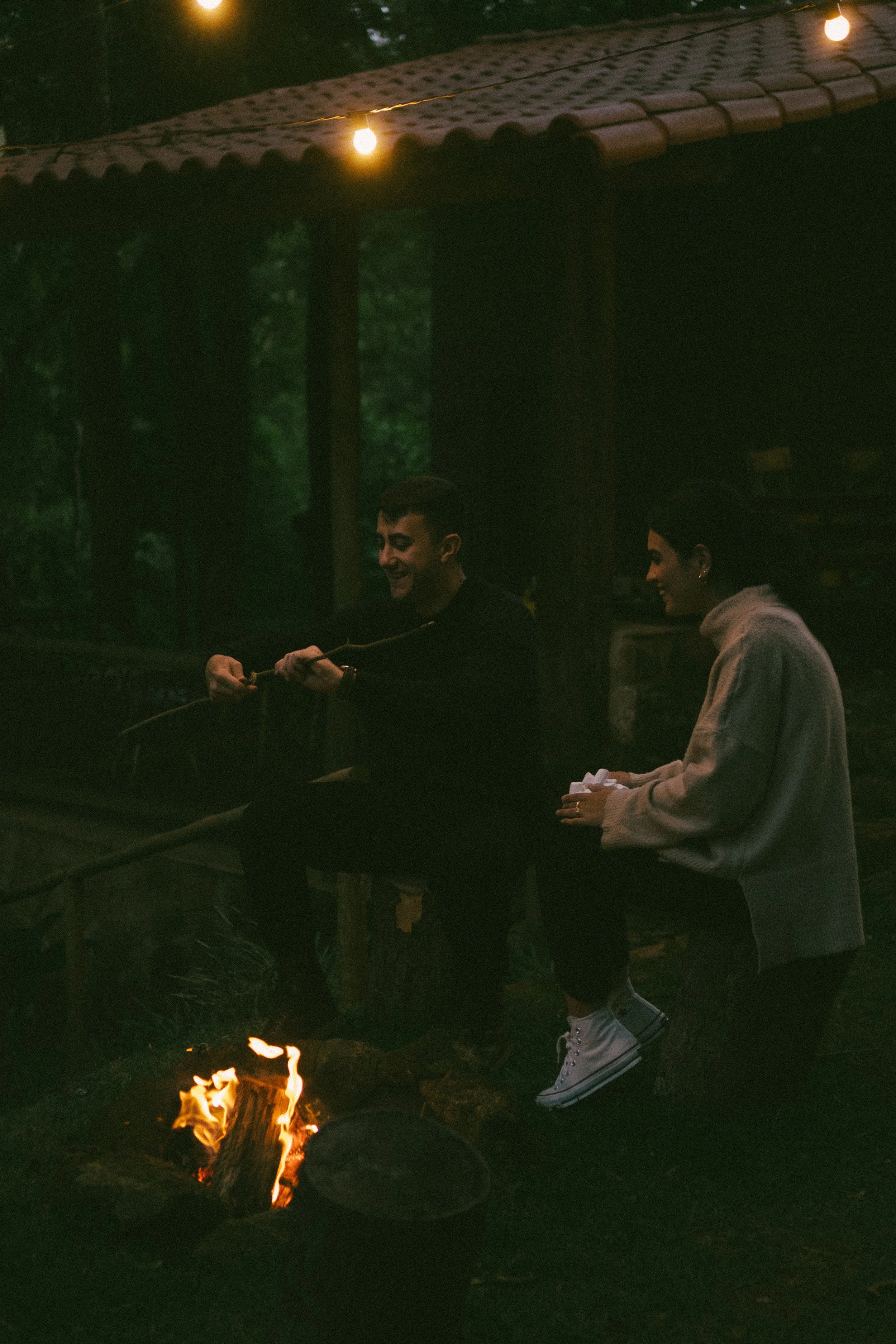 Two people sitting around a campfire at night, one is smiling while holding a stick, the other is seated with a paper towel in hand, in an outdoor setting with string lights overhead.
