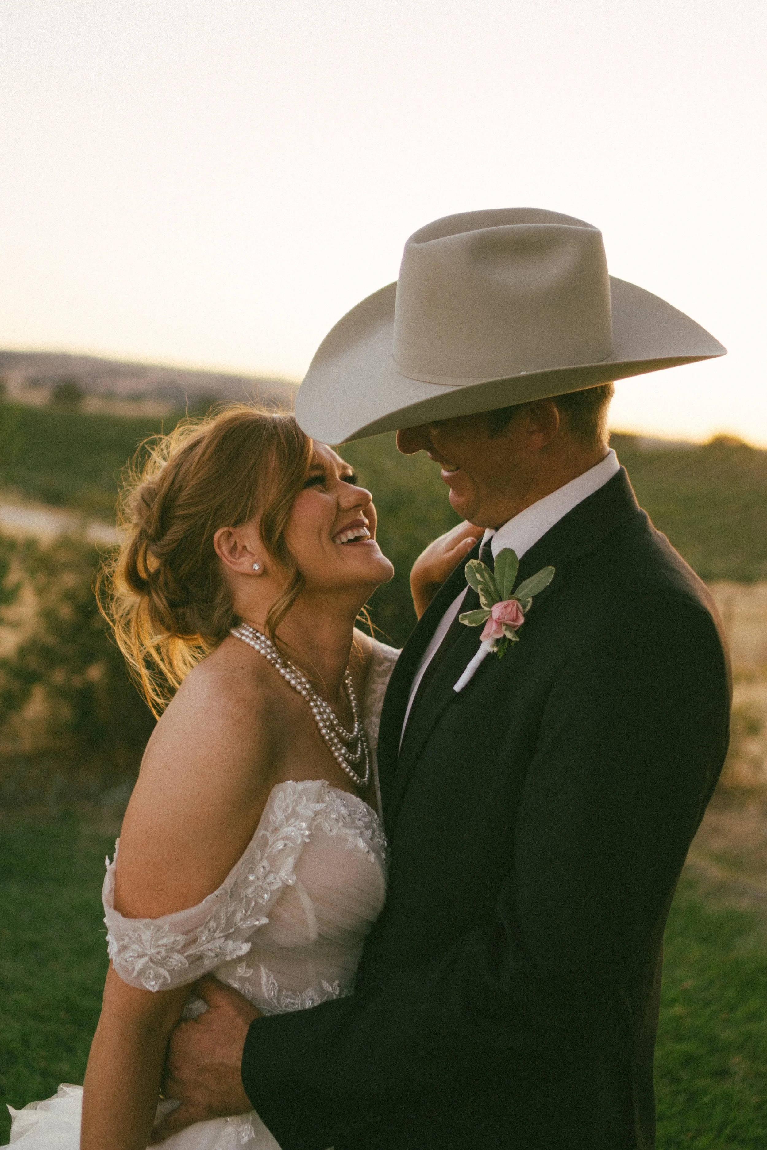 A bride and groom sharing a joyful moment outdoors at sunset, with the groom wearing a large cowboy hat and a boutonniere, and the bride wearing a lace wedding dress and pearl jewelry.
