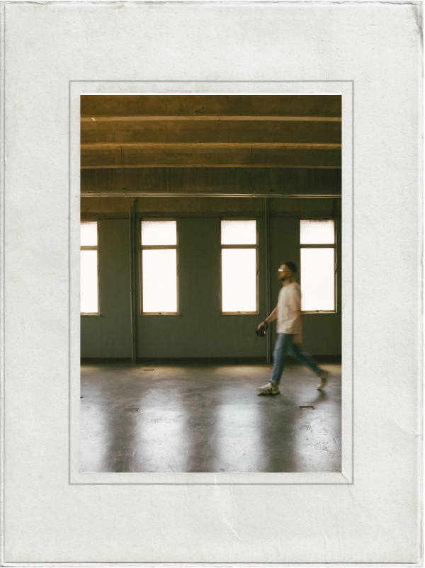 A man walking inside a room with four large windows, concrete floor, and exposed ceiling beams, holding an item in his right hand.