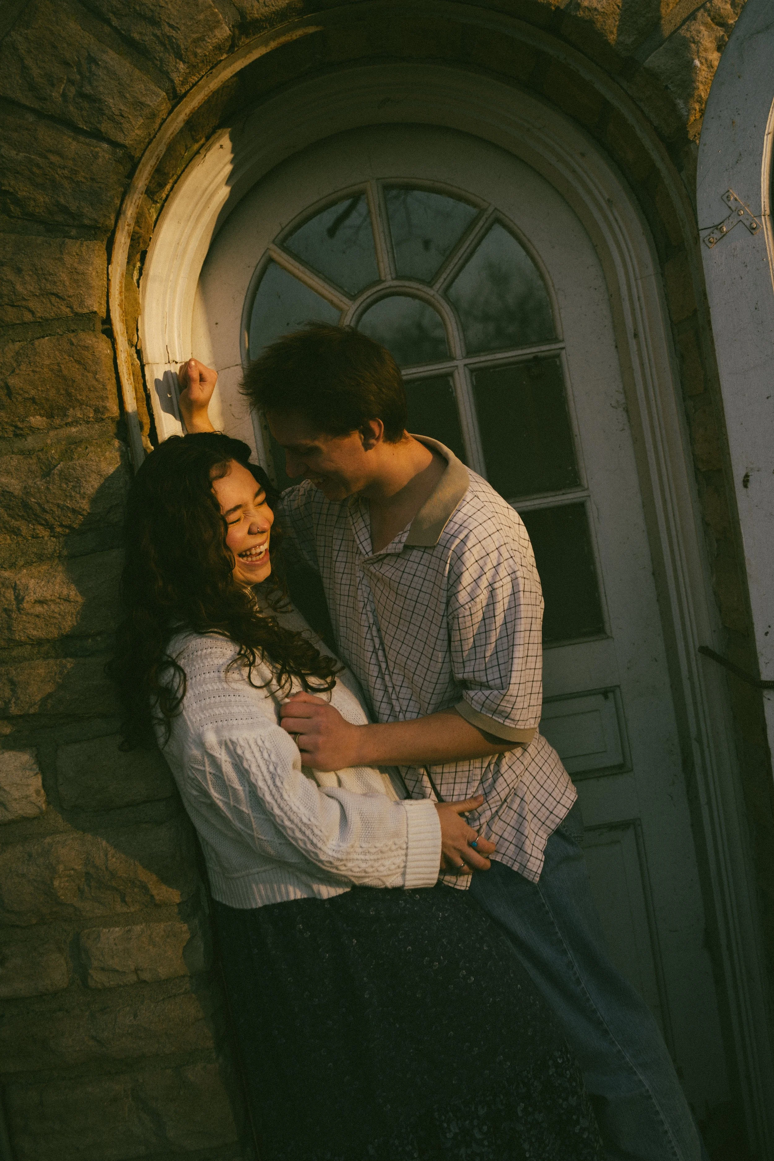 A young couple is standing close together, smiling and laughing in front of a stone wall and a white arched window.