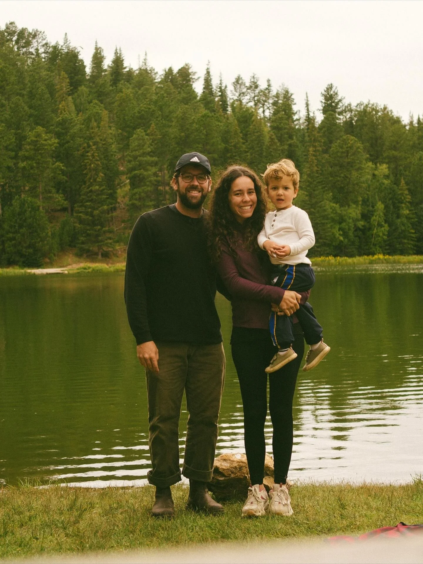 Family of three standing by a lake with trees in the background. The woman is holding a young boy, and a man is standing beside them, all smiling.