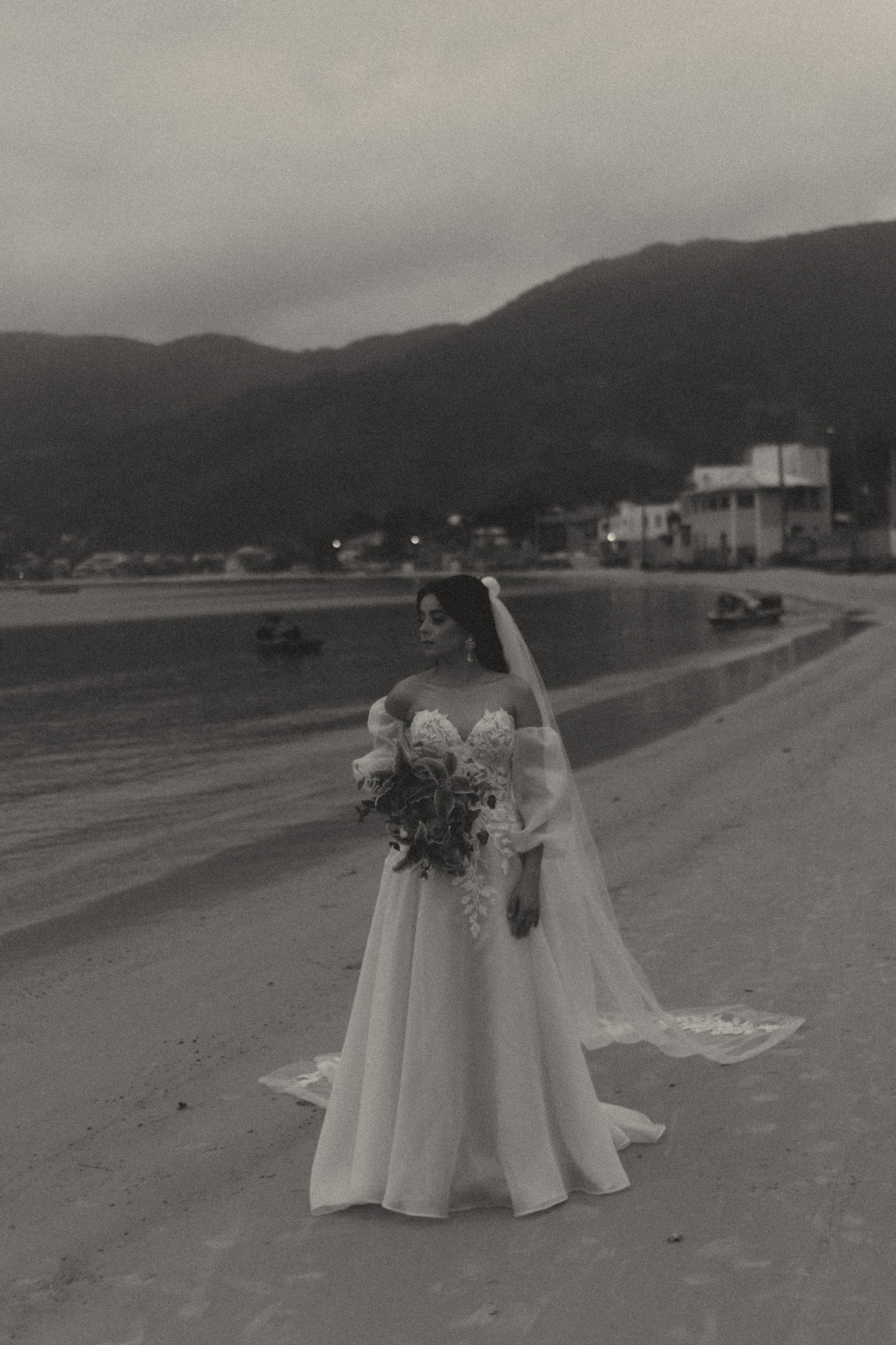 Black and white photo of a woman in a wedding dress holding a bouquet, standing on a beach with mountains and houses in the background.