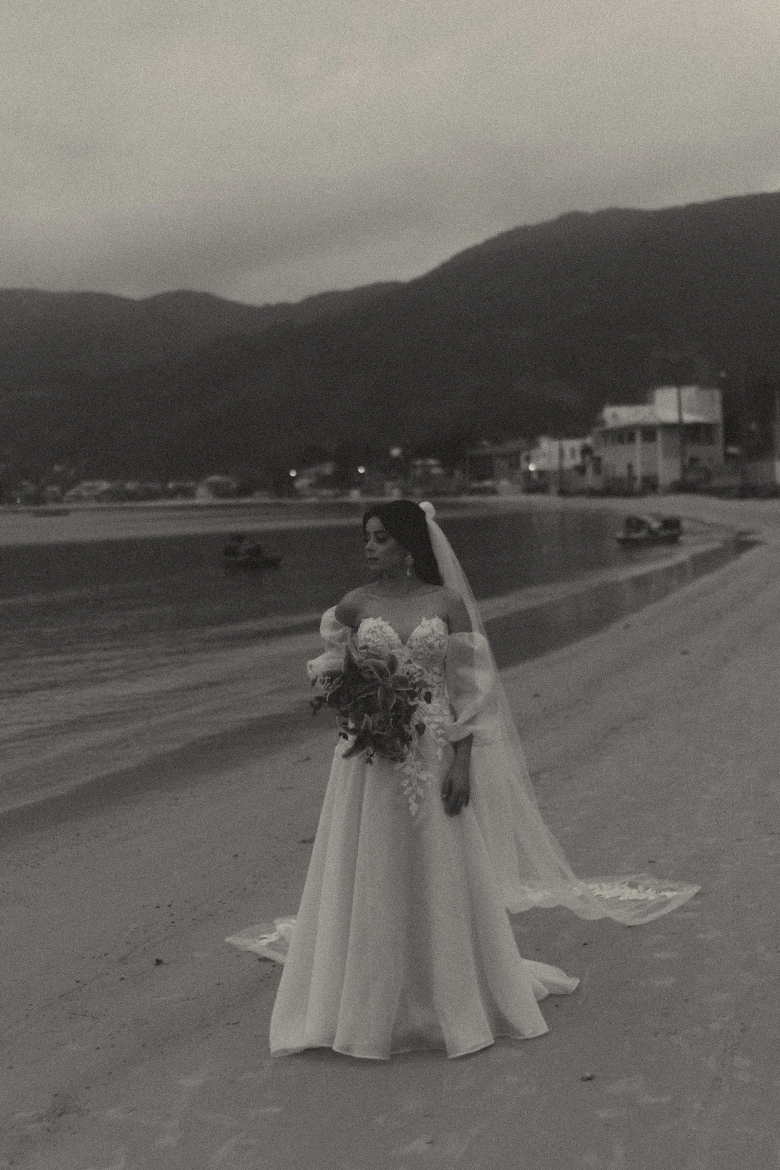 A woman in a wedding dress standing on a beach with mountains and buildings in the background, holding a bouquet.