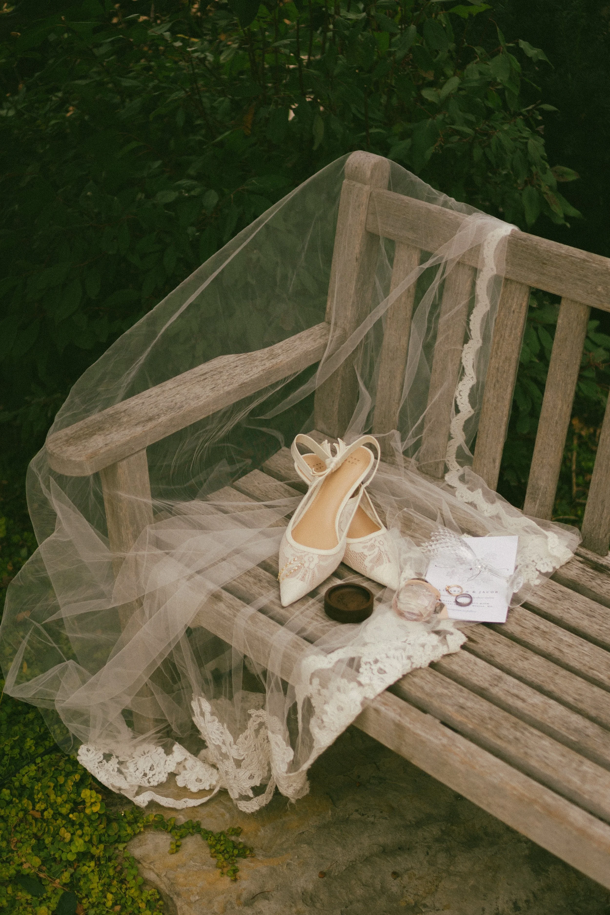A pair of wedding shoes, a veil, a ring, a card, a small box, and some perfume are placed on a wooden bench outdoors, surrounded by greenery.
