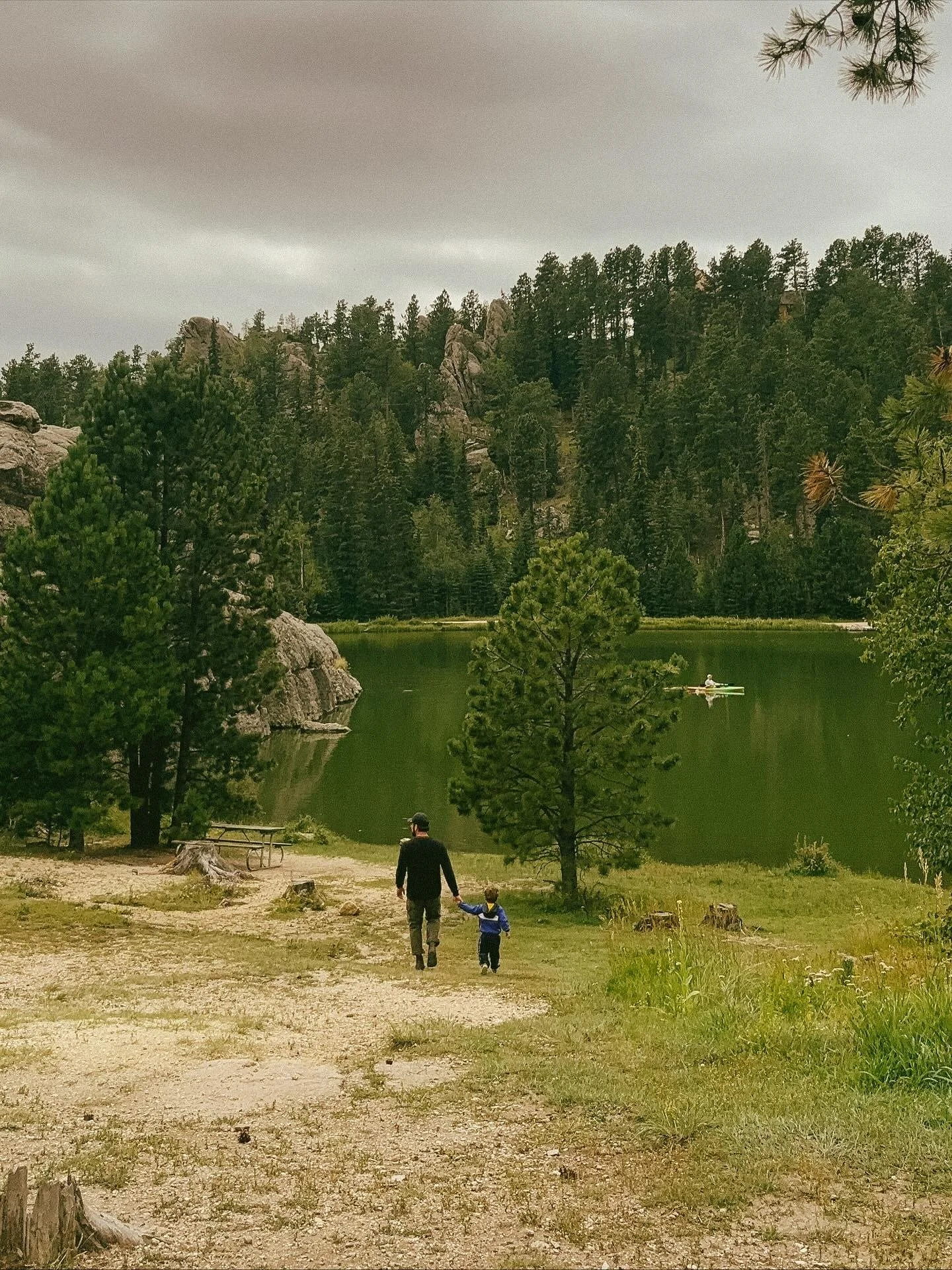 A man and a child walking on a dirt path next to a lake, surrounded by trees and mountains, with a kayaker on the water.