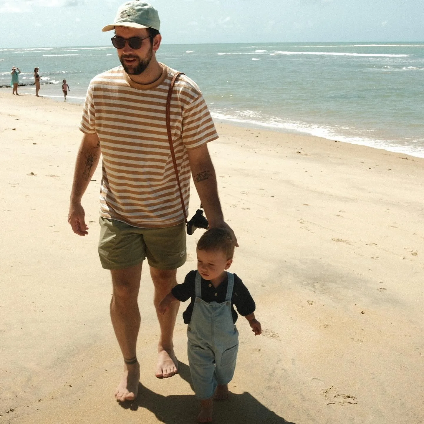 A man and a young boy walking on a sandy beach near the ocean, with other people in the background. The man is wearing a striped t-shirt, shorts, and a cap, while the boy is dressed in overalls and a dark shirt.