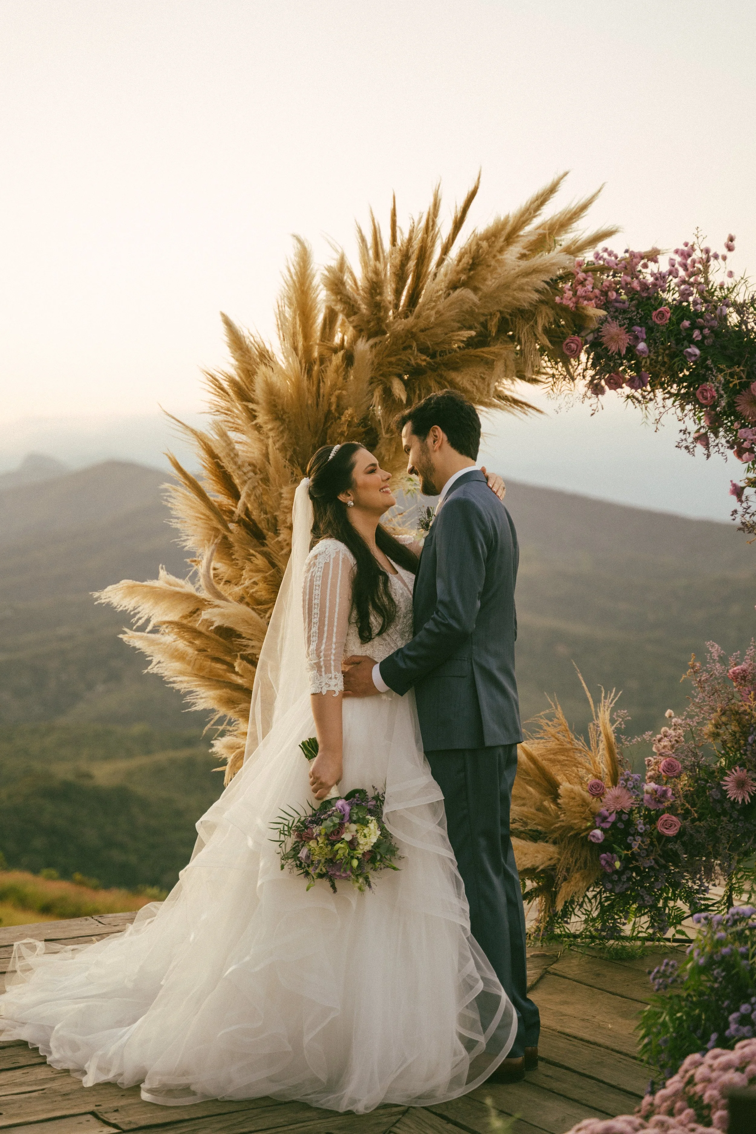 A bride and groom embrace during their outdoor wedding ceremony, standing on a wooden platform with a scenic mountainous background, floral decorations, and pampas grass arch.