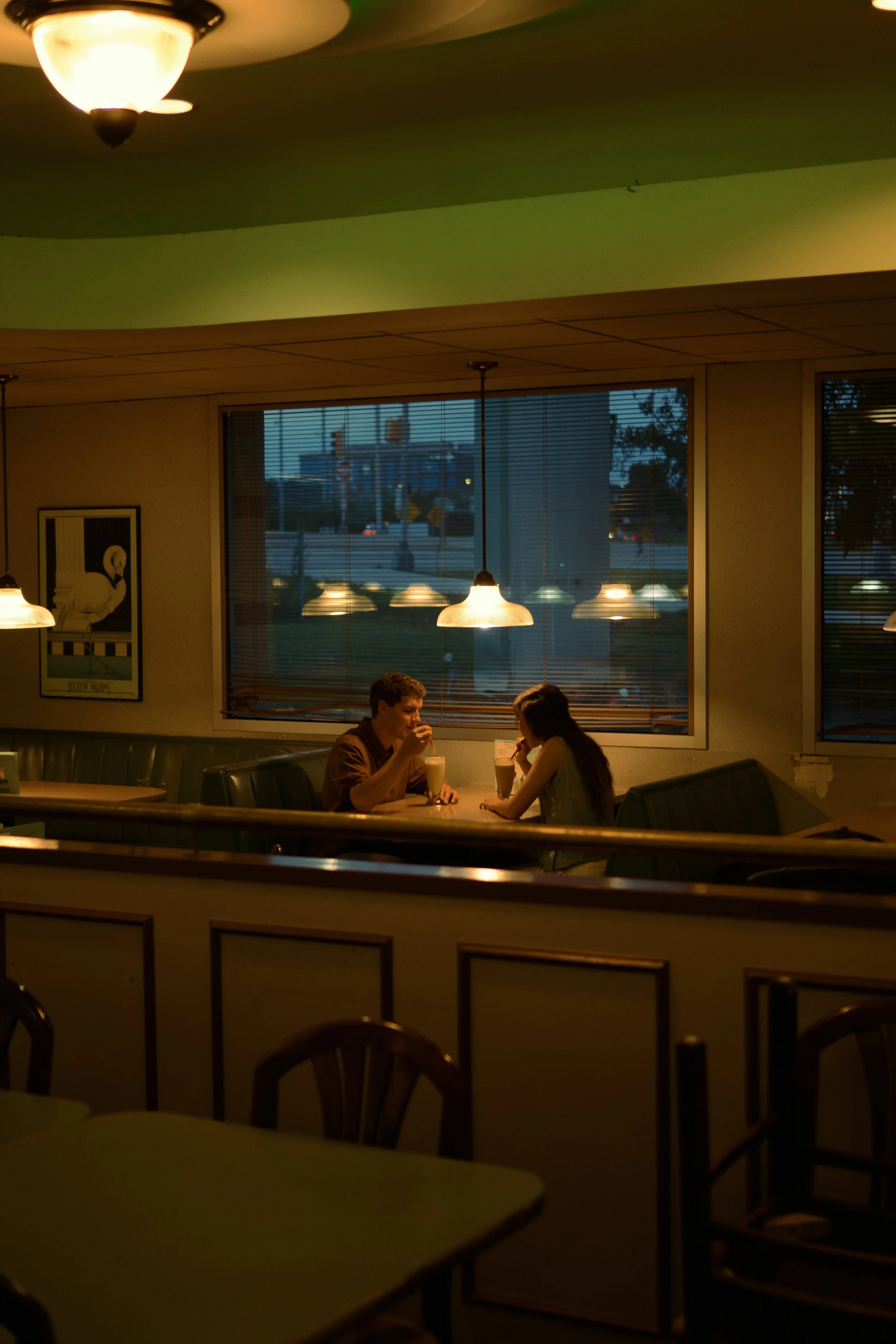 Two people sitting at a table in a dimly lit diner, drinking milkshakes, with large windows showing a city street outside at dusk.