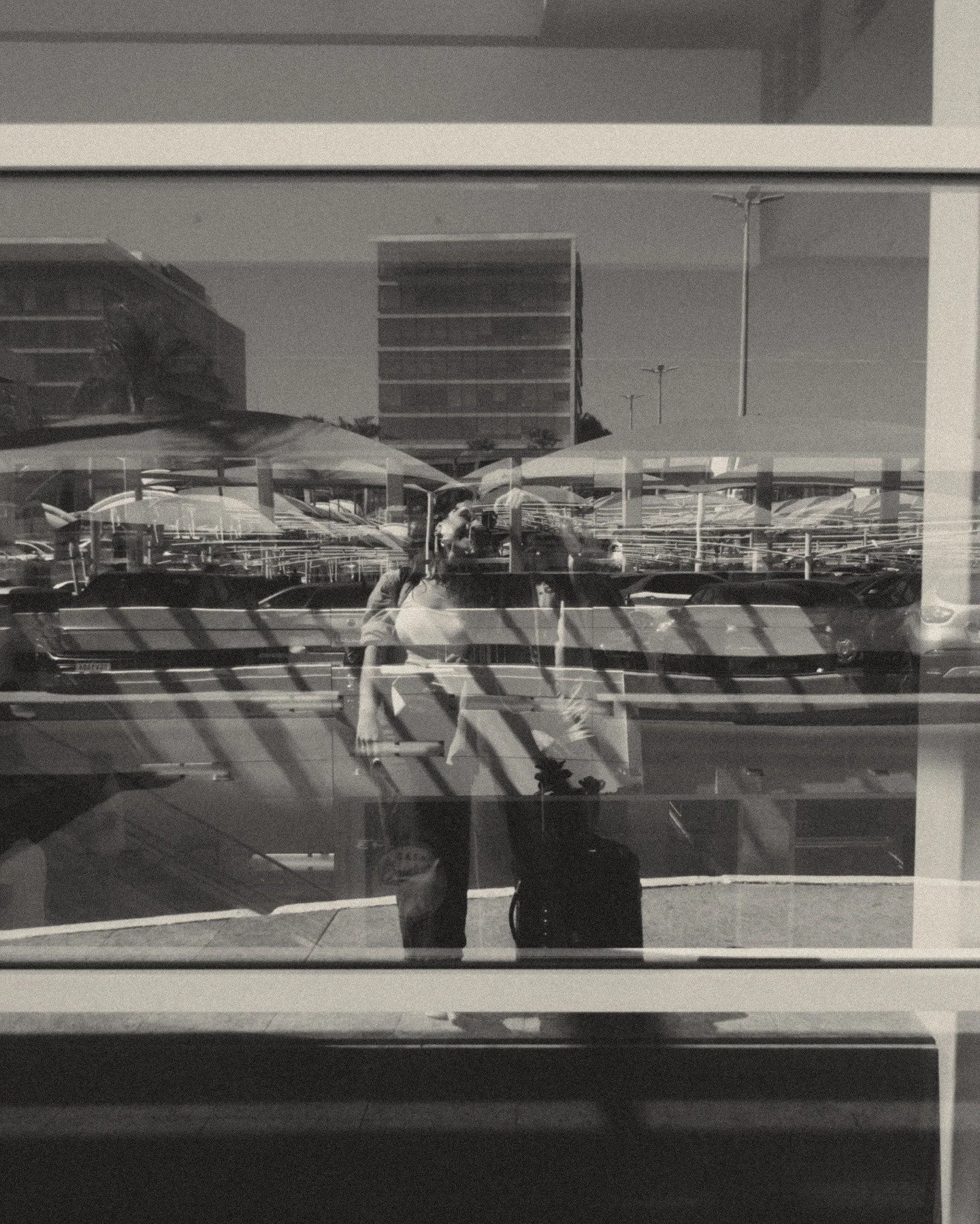 Reflections of two women with bags and a person in a parking lot, seen through a window with horizontal blinds, with parked cars and multi-story buildings in the background.