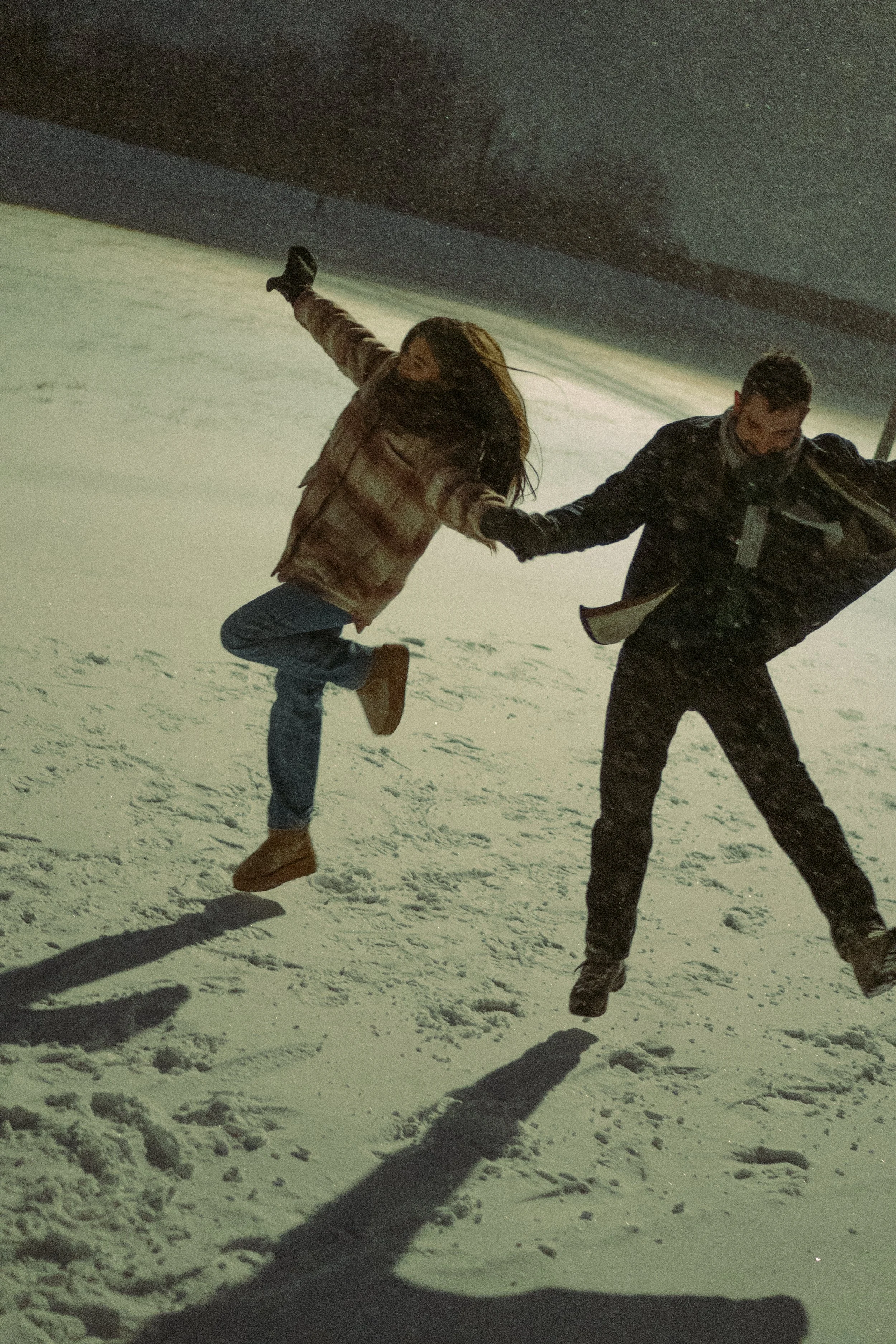 Two people holding hands and spinning in the snow at night, with their shadows cast on the snow and a dark sky overhead.