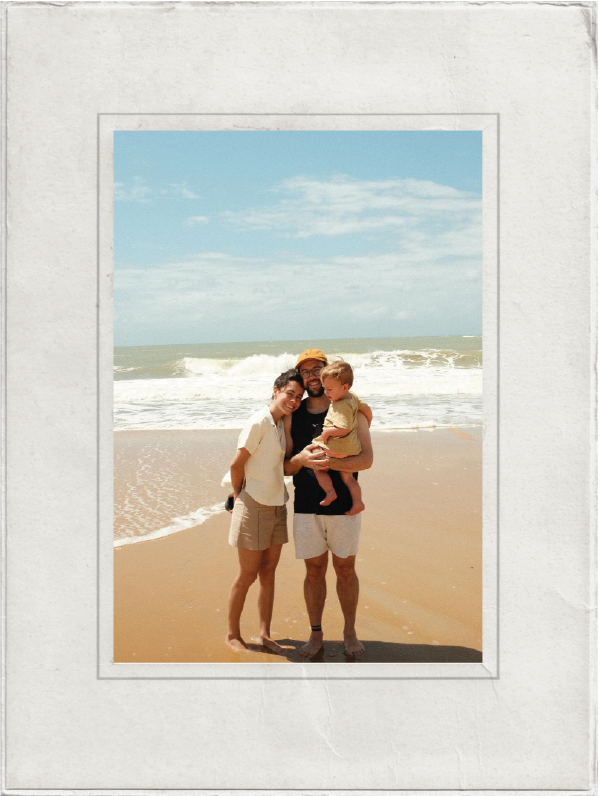 A family of three standing on a sandy beach with ocean waves in the background. The man is wearing a hat and sunglasses, holding a young child. The woman is standing close, smiling, with the child in the man's arms. The sky is partly cloudy.