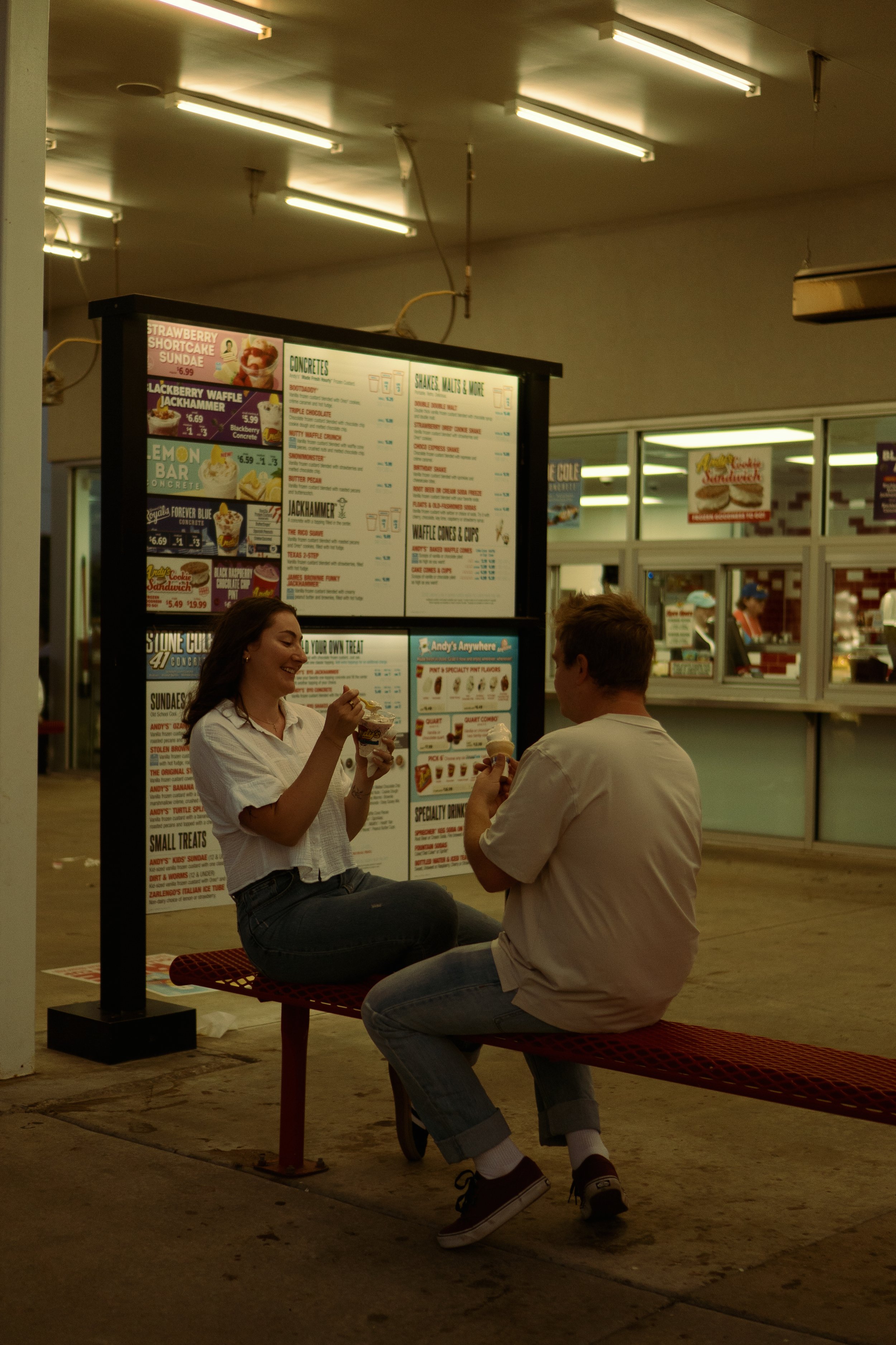 A young woman and a young man sitting on a red bench outside a fast-food restaurant, eating ice cream and engaging in conversation. The scene is illuminated by overhead fluorescent lights, with a large menu board in the background.