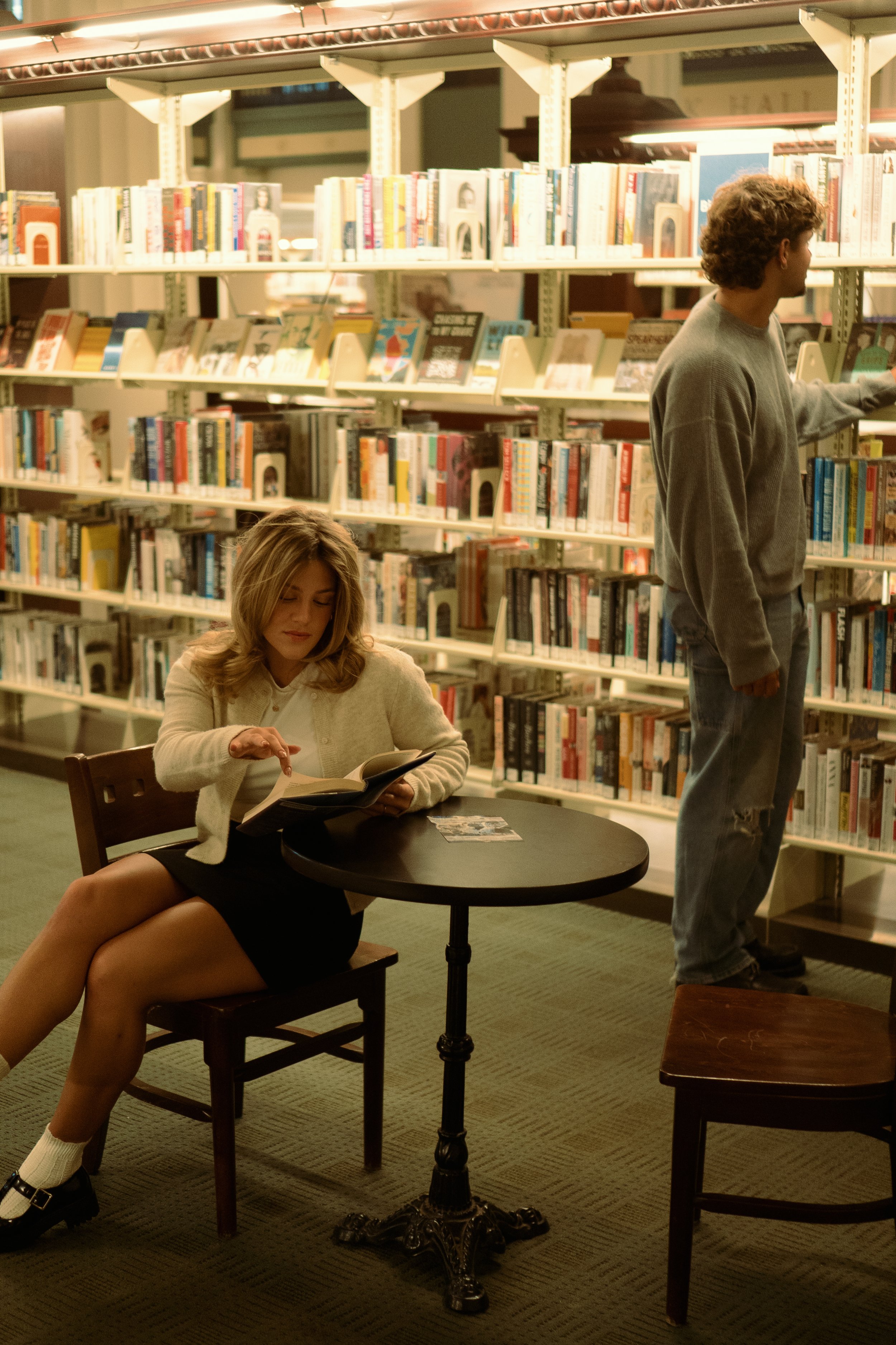 A woman sitting at a small table in a library, reading a book, with a man browsing bookshelves nearby.