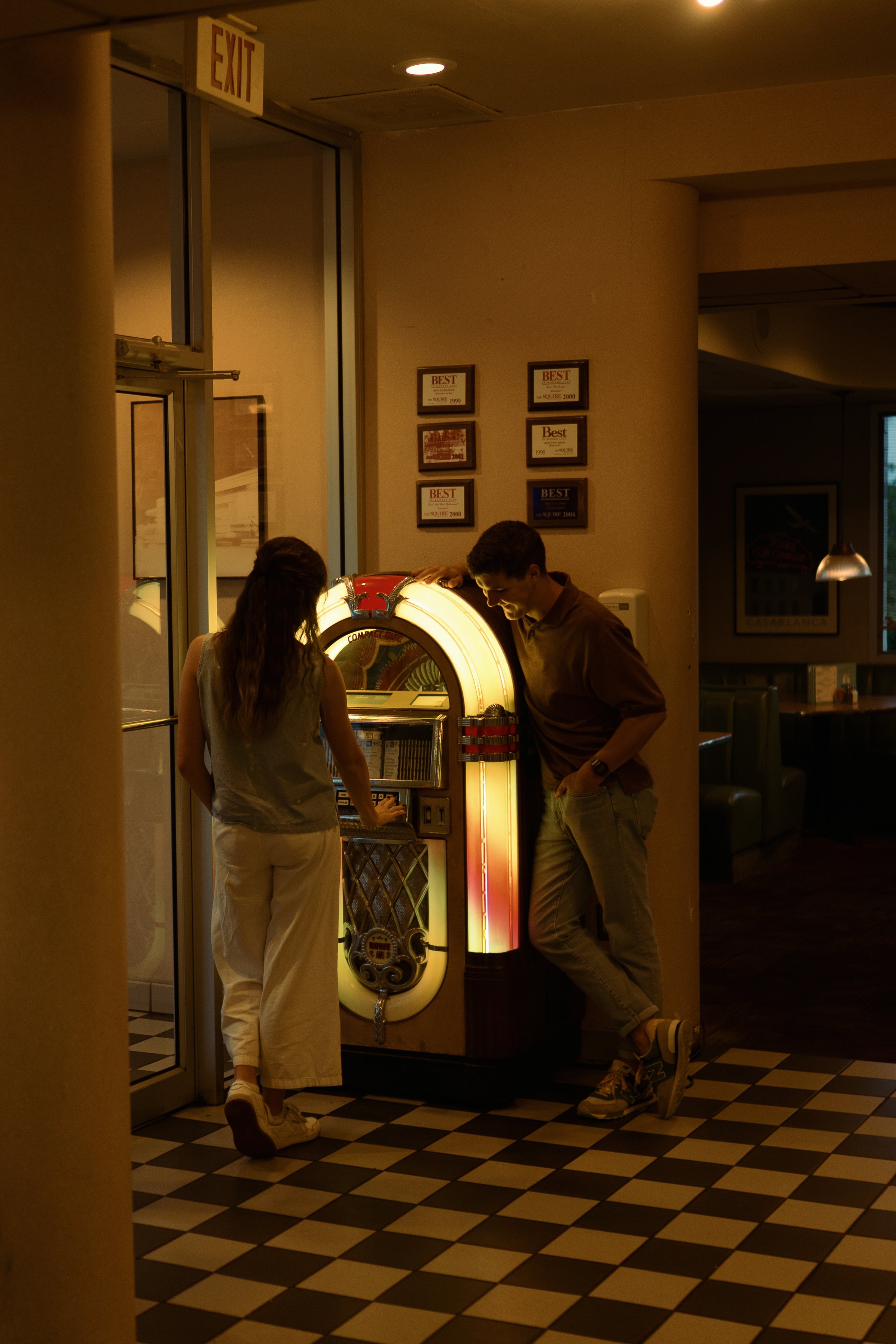 Two young people, a woman and a man, standing next to a vintage jukebox in a dimly-lit restaurant or diner. The woman is pressing buttons on the jukebox while the man leans against it, smiling and wearing casual clothing.