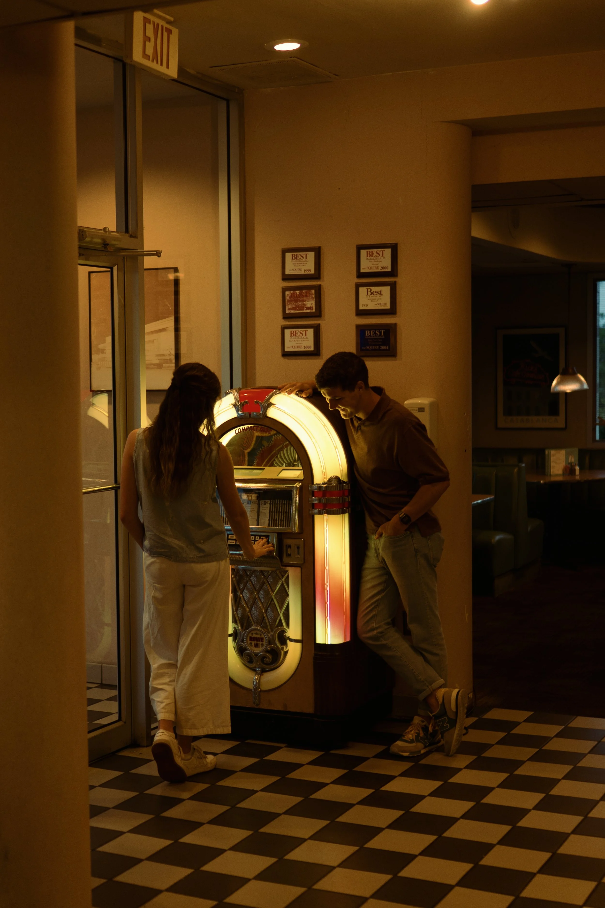 Two young people, a woman and a man, standing next to a vintage jukebox in a dimly-lit restaurant or diner. The woman is pressing buttons on the jukebox while the man leans against it, smiling and wearing casual clothing.