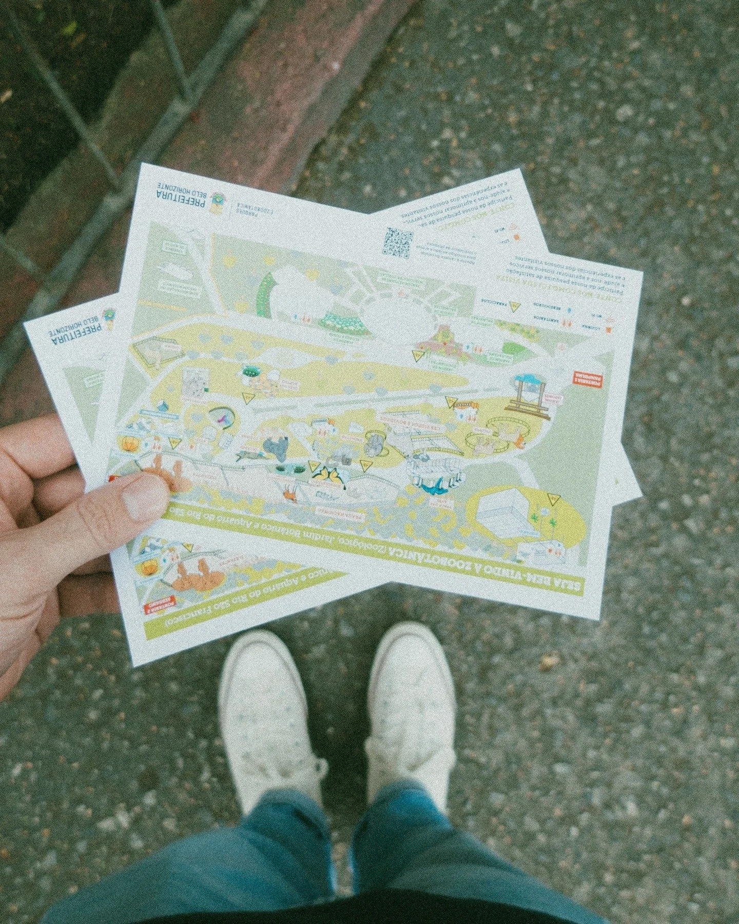 Person holding a map or brochure, standing on a sidewalk wearing white sneakers and blue jeans.