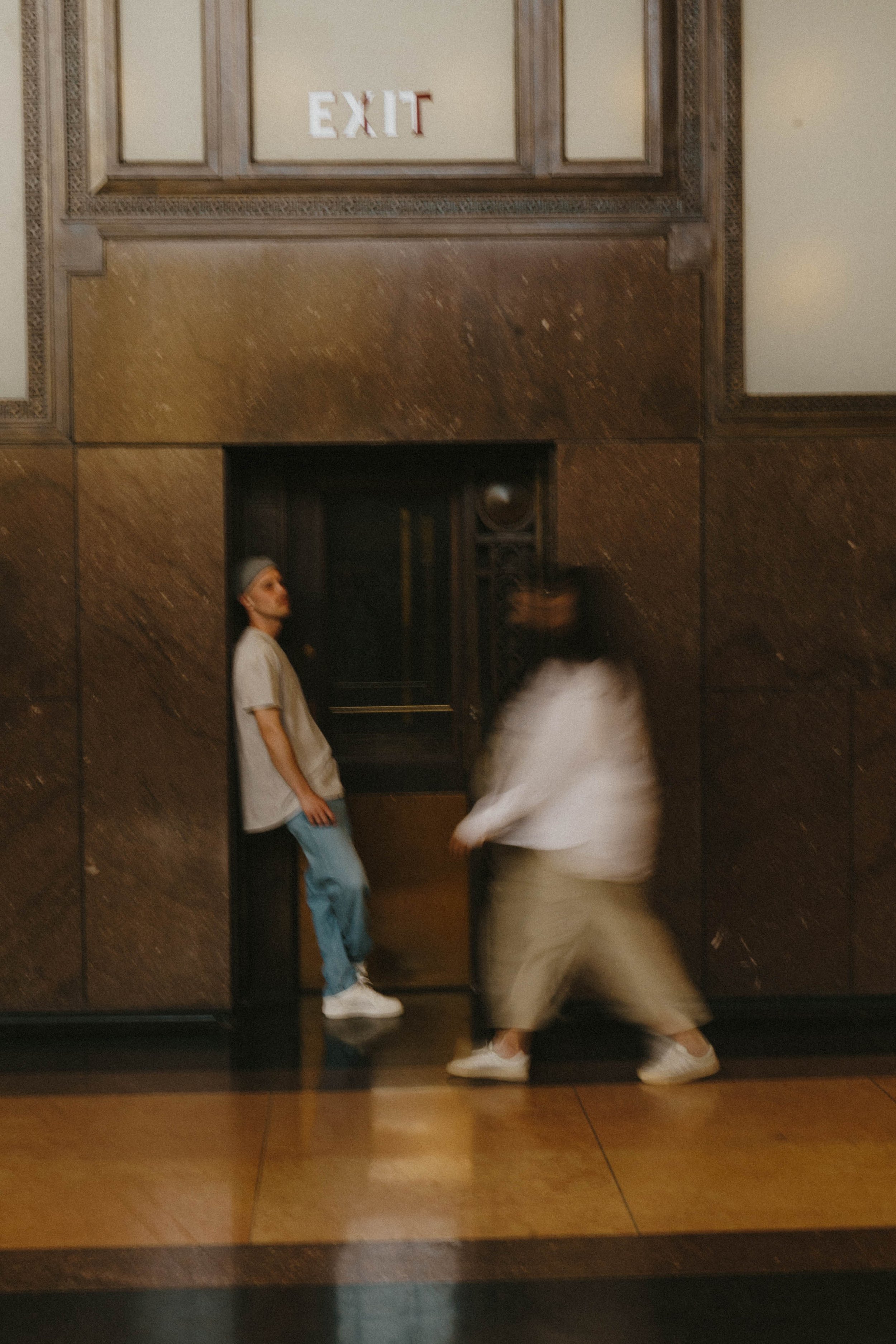 Two people are near an elevator door; one person is standing against the wall, and the other is walking past.