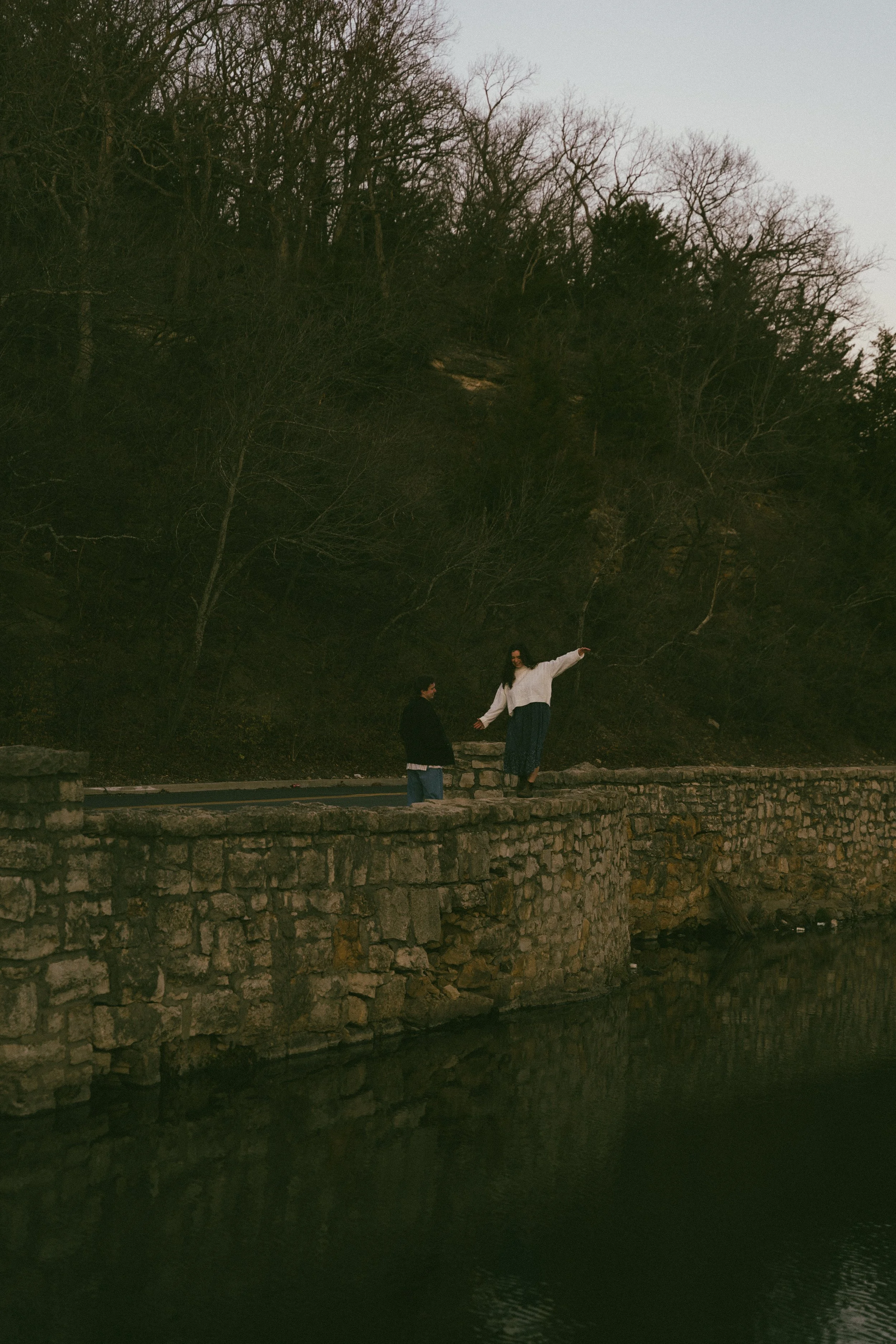 A woman standing on a stone wall near a body of water, with her arms extended, and a man standing nearby, against a backdrop of leafless trees on a hillside during dusk.