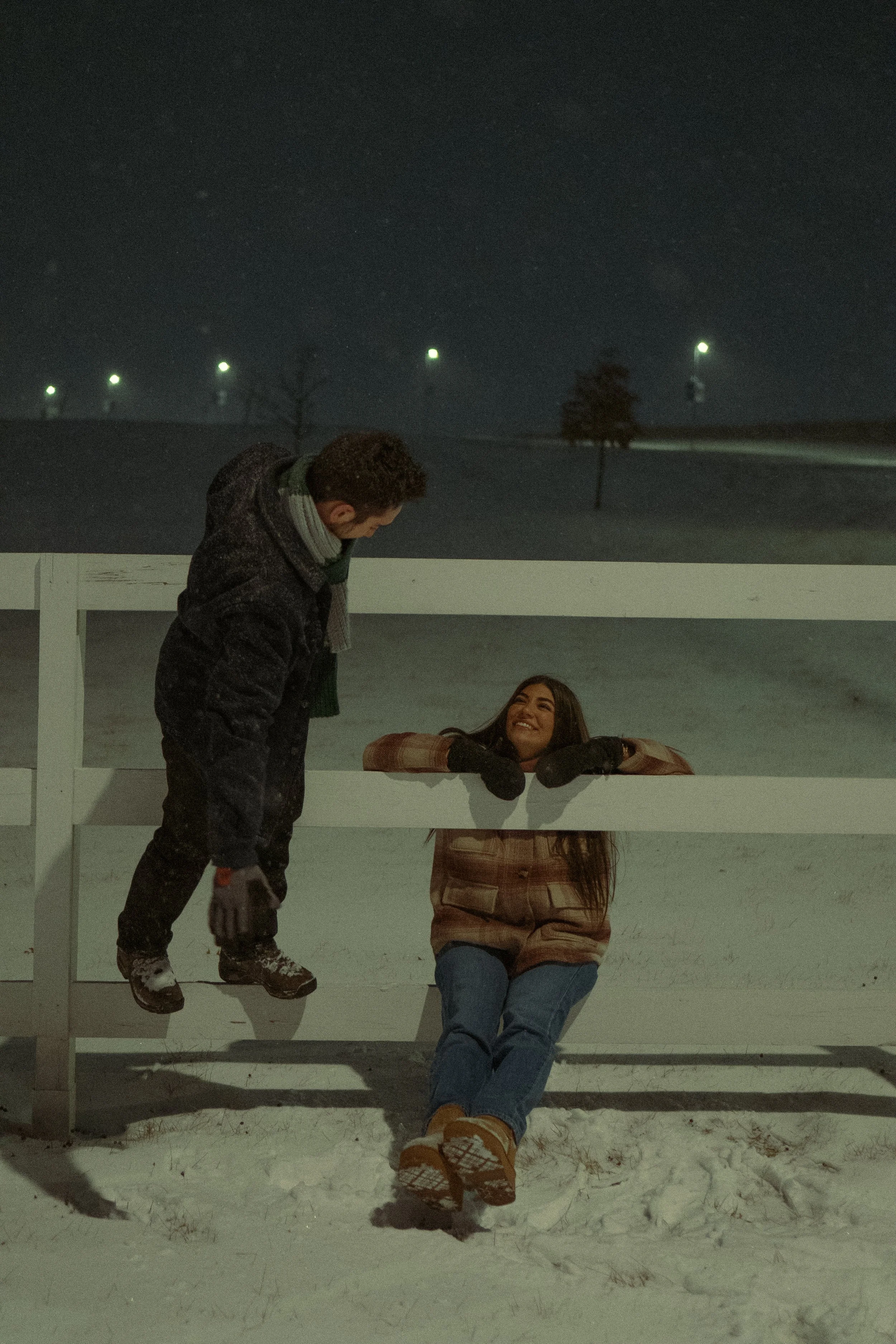 A man leaning over a girl sitting on the snow in a winter landscape at night, with trees and distant streetlights in the background.