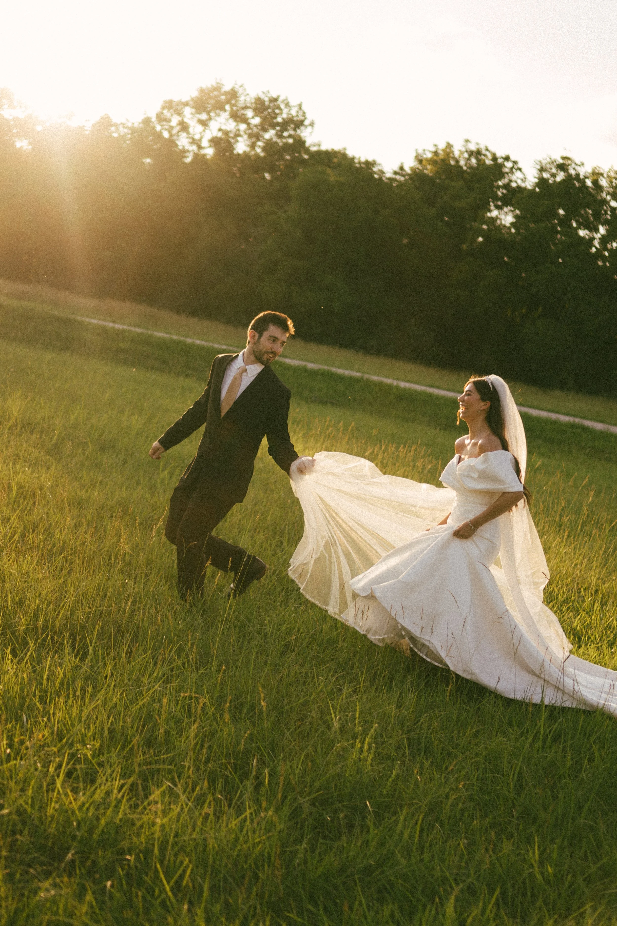A bride and groom dancing in a grassy field during sunset, with the groom holding the bride's dress.