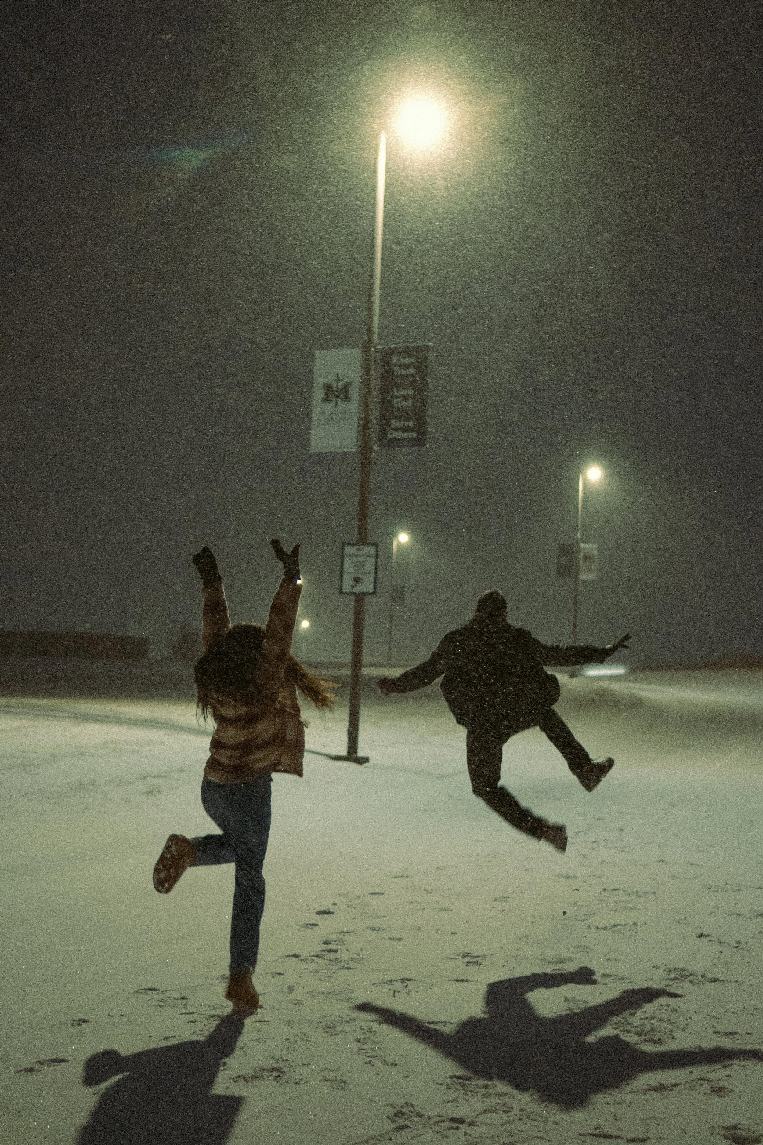 Two people are jumping and playing in a snowy, nighttime outdoor scene illuminated by streetlights. One person is raising their arms with enthusiasm while the other swings their leg mid-air. Snow is falling, creating a lively winter atmosphere.