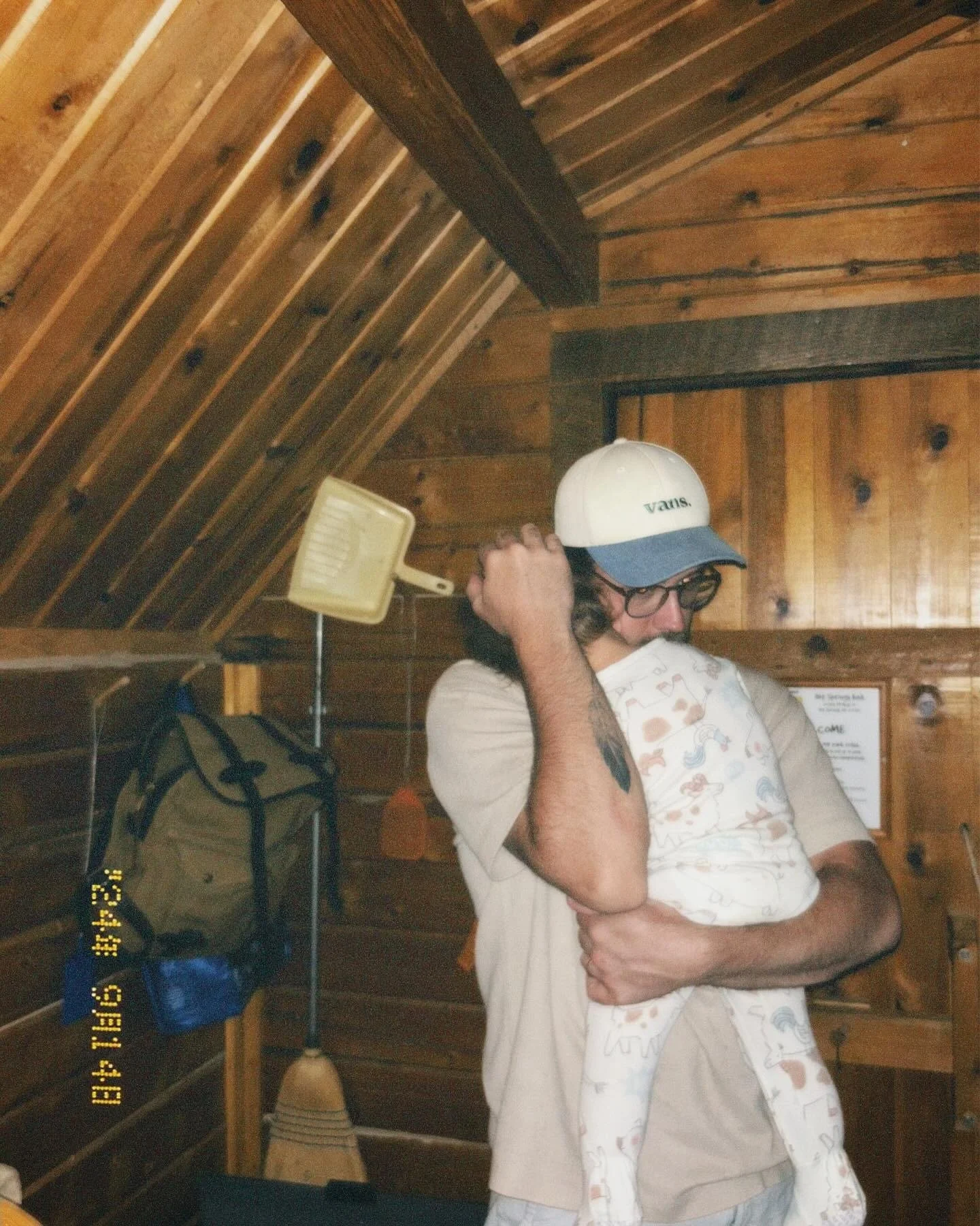 A man holding a child inside a wooden cabin, with wooden walls and ceiling. The man is wearing glasses and a white cap with a blue brim, and the child is wearing a white top with colorful animal prints.