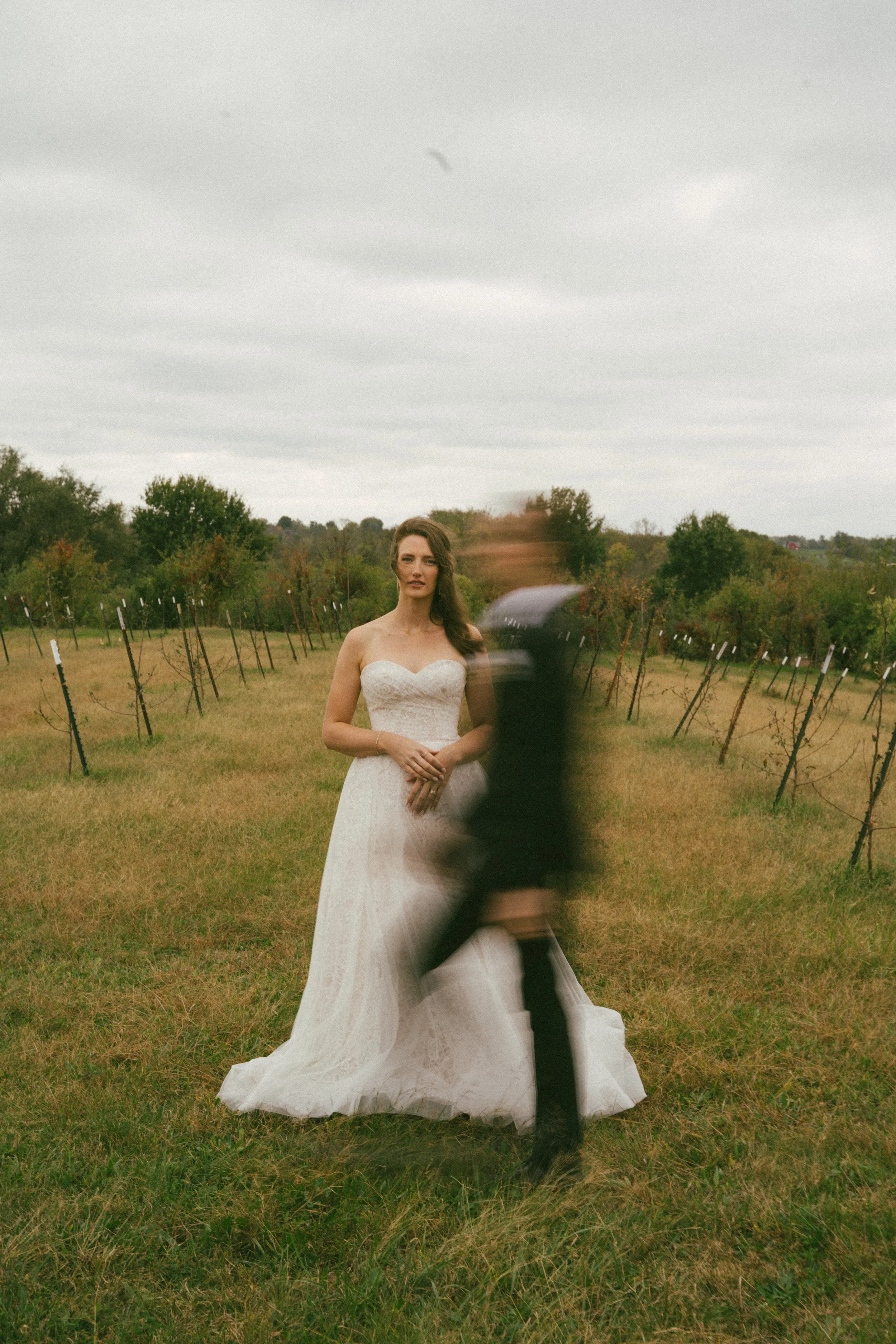 A bride in a white wedding dress standing in a vineyard with blurred man walking in front of her on a cloudy day.