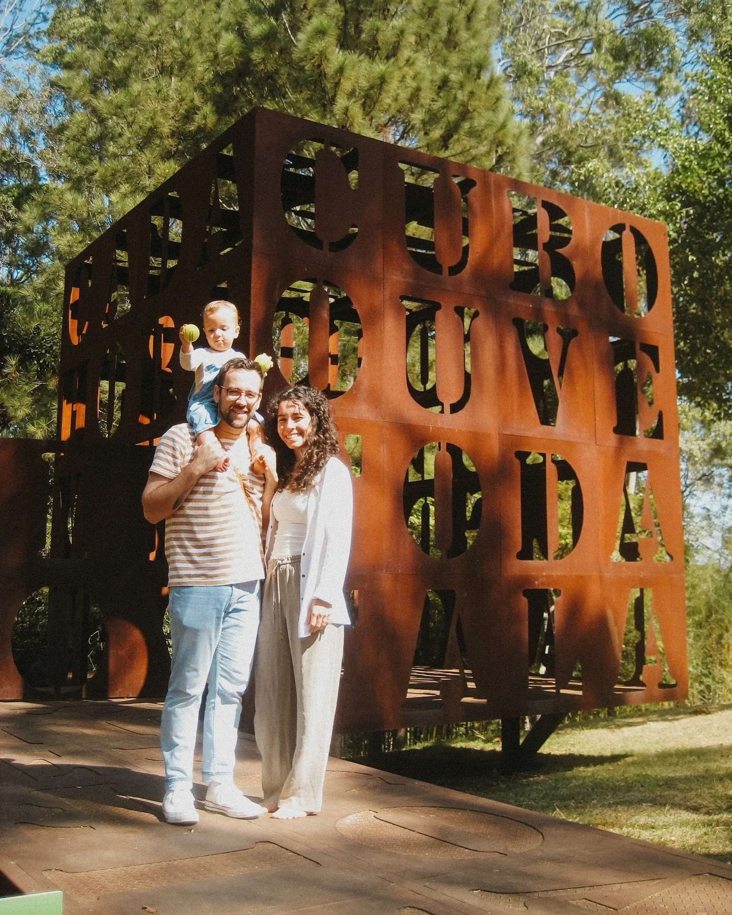A family of three standing in front of a large, rust-colored metal sculpture. The sculpture spells out the word 'COURAGE' in large letters with the phrase 'to love, to forgive' underneath. The child is sitting on the man's shoulders, holding what appears to be two tennis balls. The family is outdoors with trees and a clear blue sky in the background.