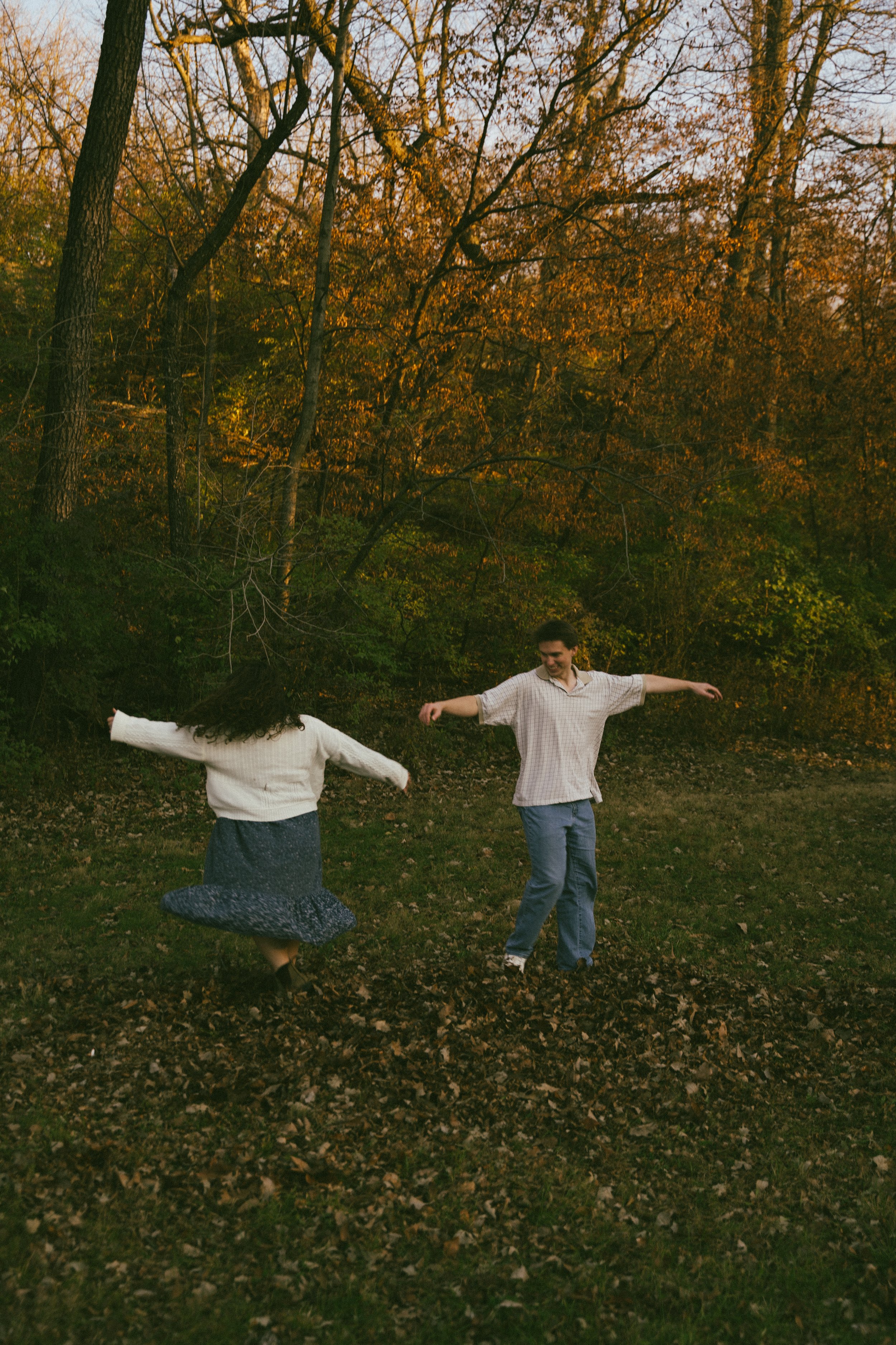 Two people dancing joyfully outdoors in a wooded area during autumn, with colorful fall foliage in the background.