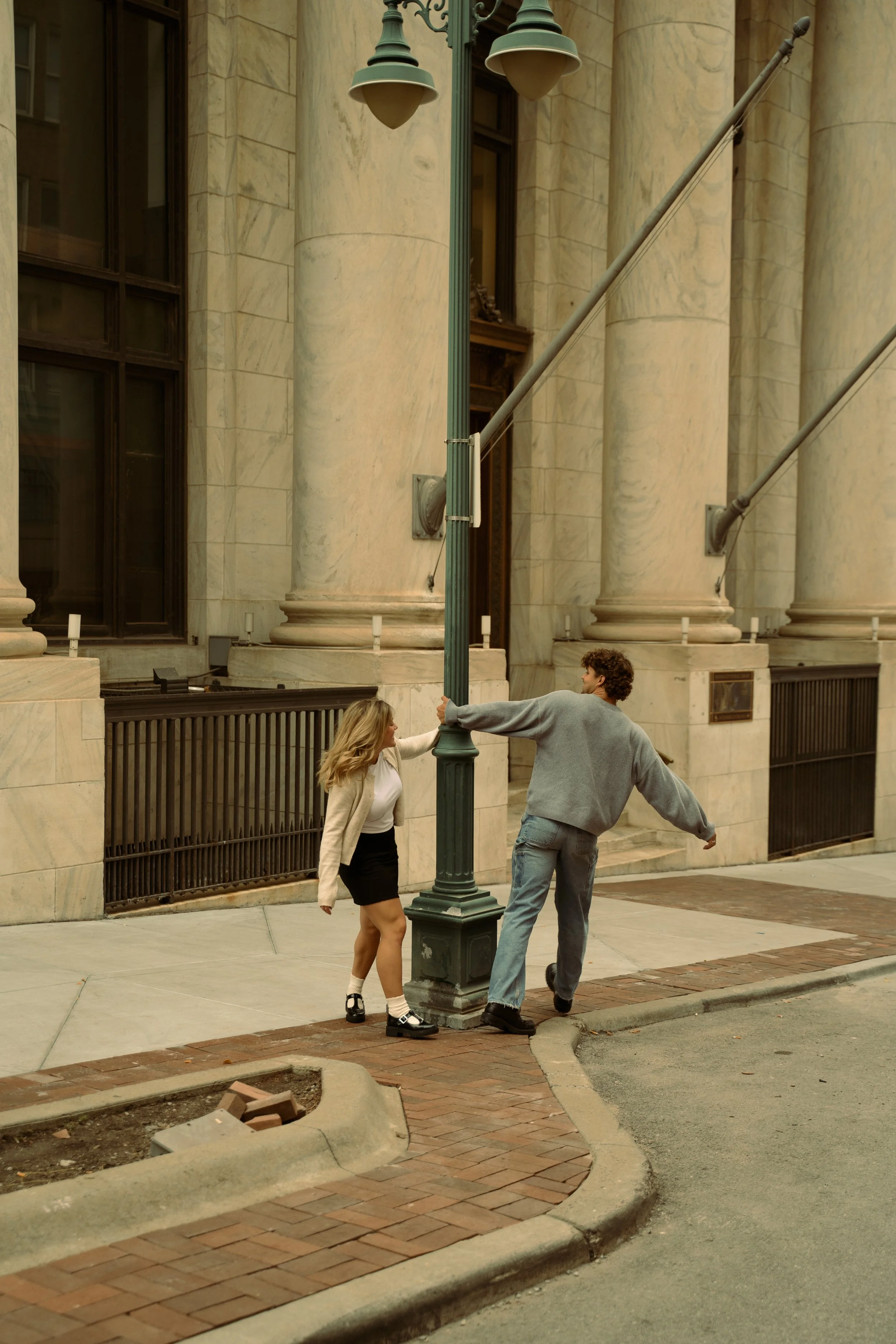 A young woman and a young man are outside, with the woman pushing the lamp post while the man holds onto it. The setting is in front of a large historic building with marble columns.