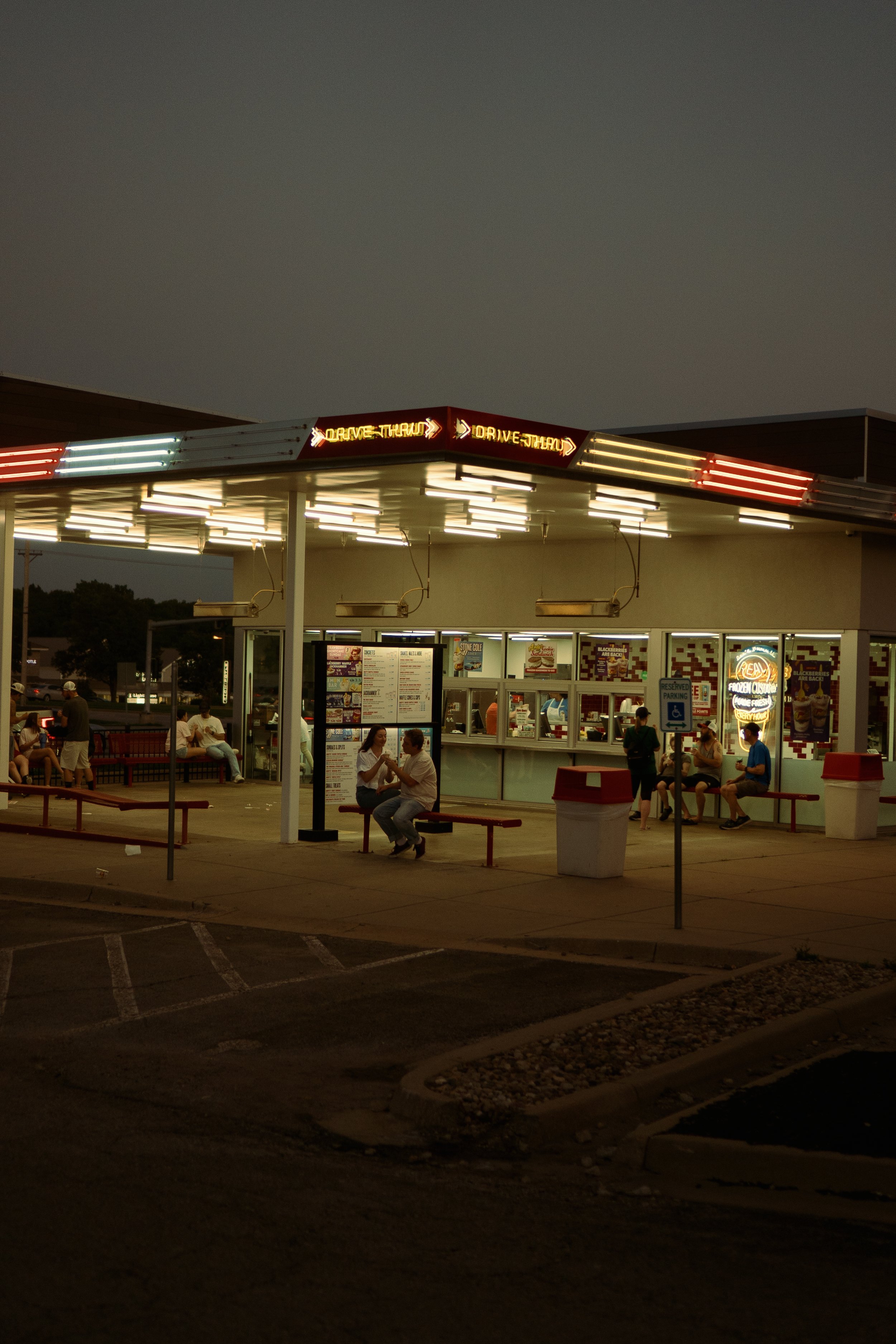 Nighttime view of a Shake Shack restaurant with outdoor seating, illuminated signs, and a small crowd of people eating and waiting outside.