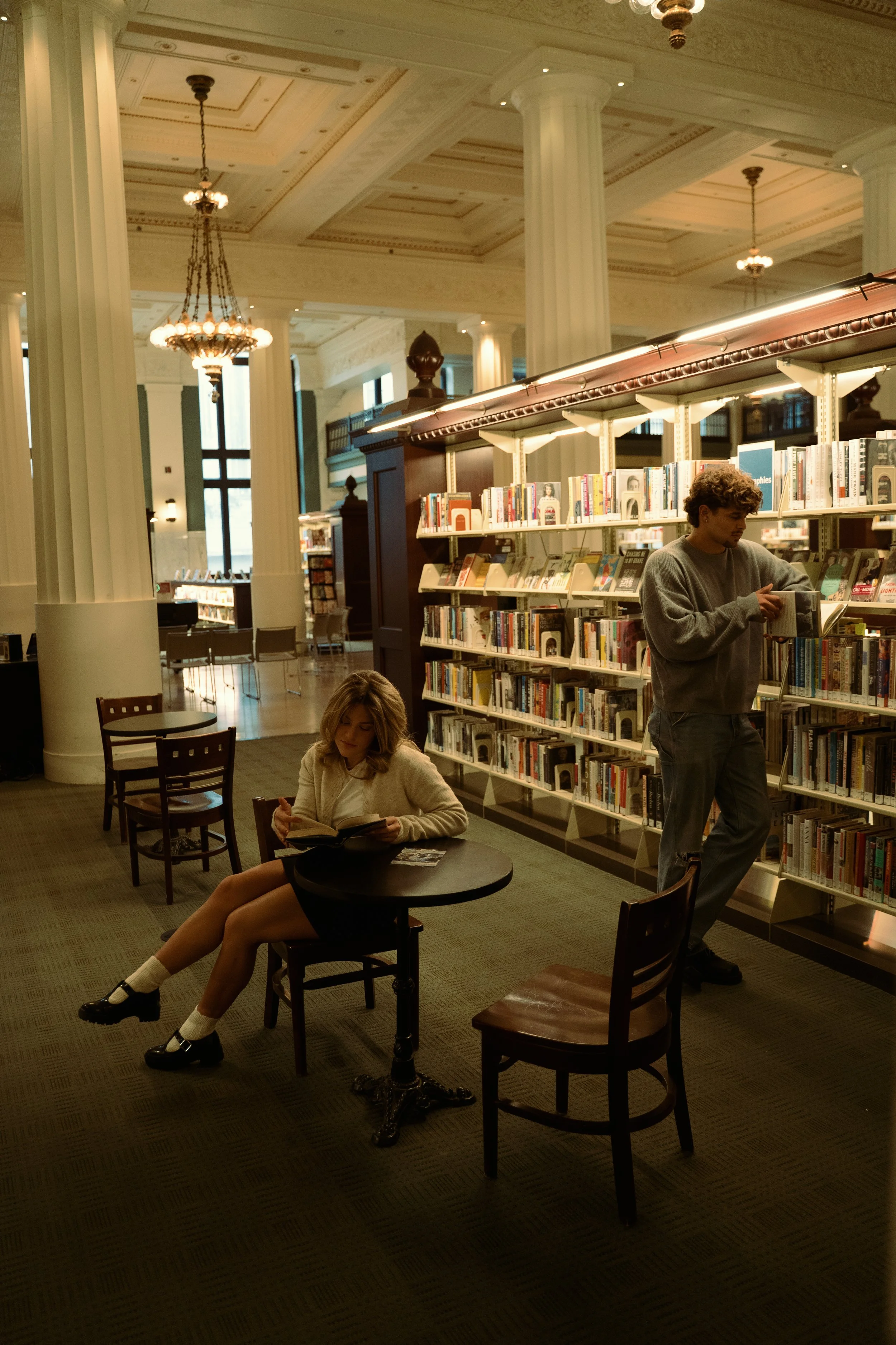 A woman sitting at a small round table reading a book and a man standing nearby browsing bookshelves inside a grand historic library.