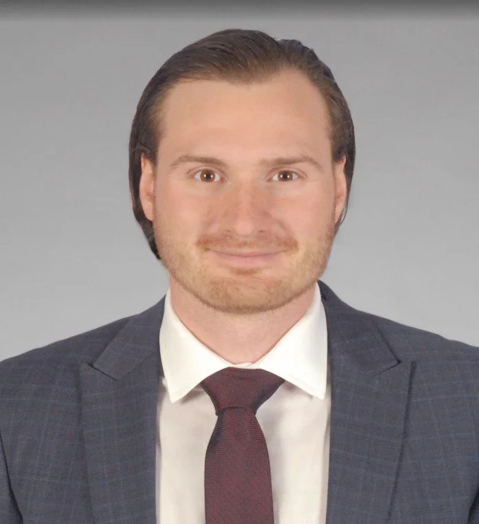 Professional headshot of a man in a suit with a white shirt and maroon tie, smiling against a plain gray background.