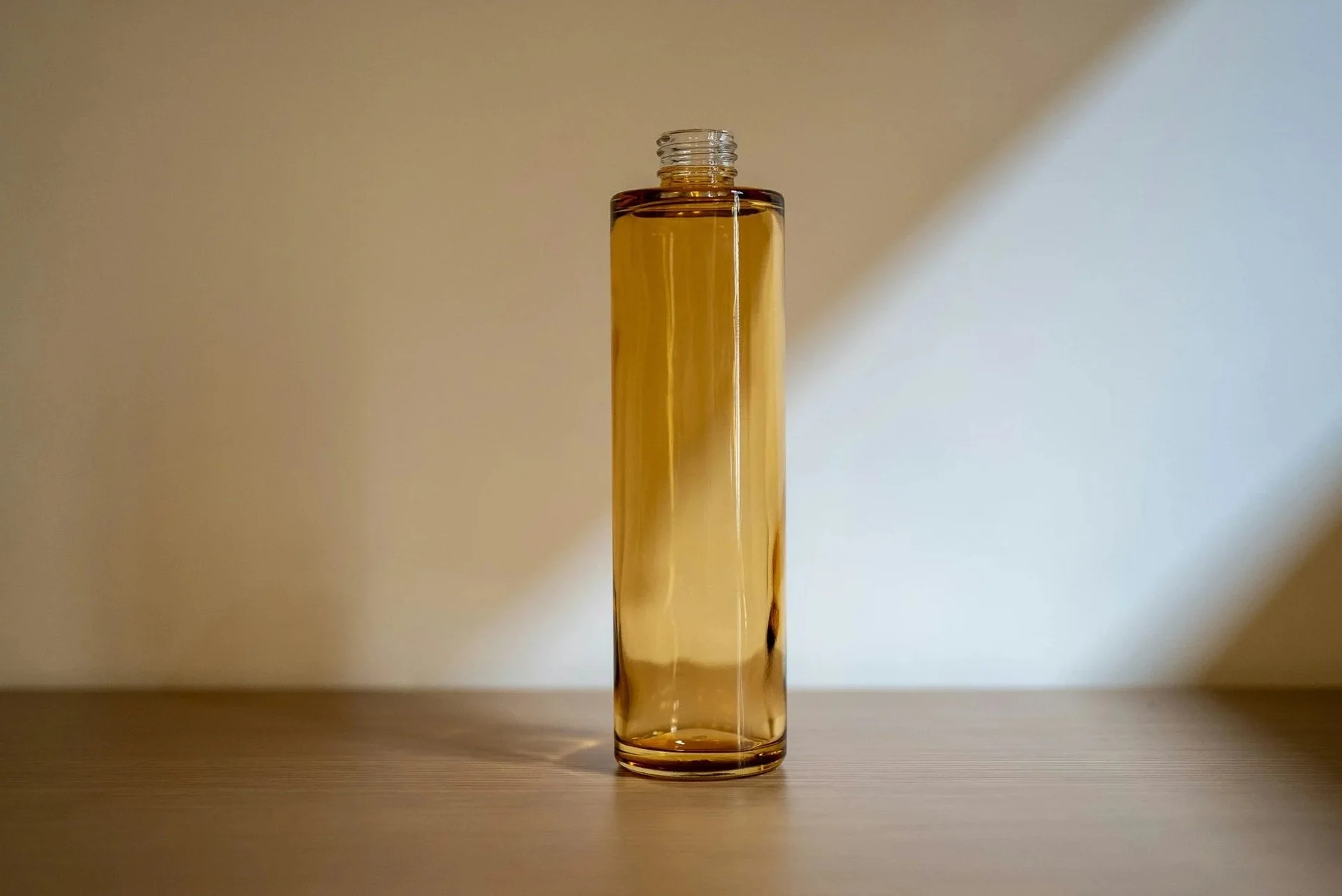 A tall, cylindrical glass bottle filled with a golden-colored liquid, placed on a wooden surface with shadow and light patterns on the plain wall behind.