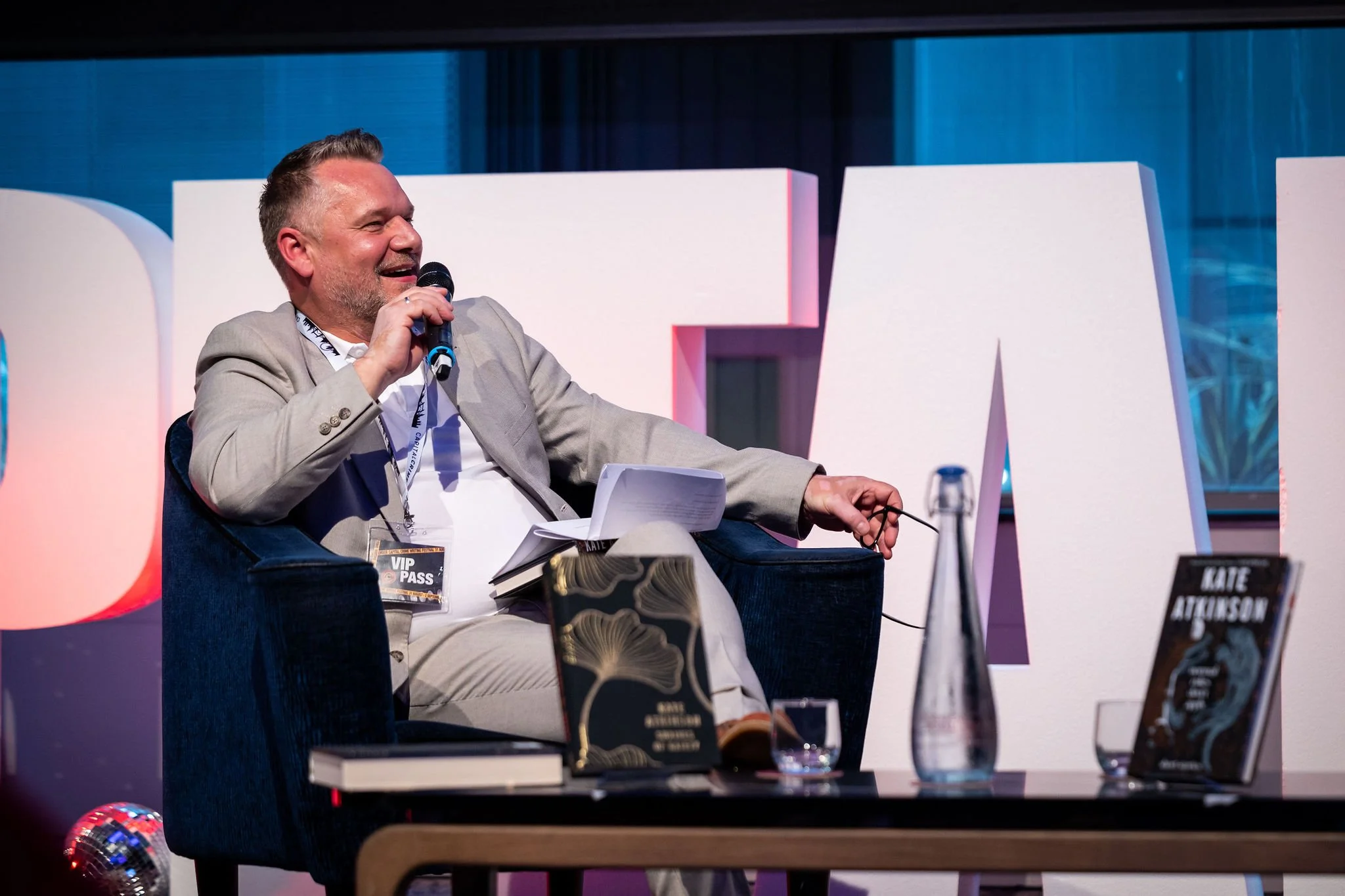 A man in a light gray suit sitting in a dark blue armchair at a book event, speaking into a microphone. There are books on the table in front of him, including one titled 'GIRLS' and another with an abstract cover. Large white letters spell out part of a word in the background, and a water bottle and glasses are on the table.