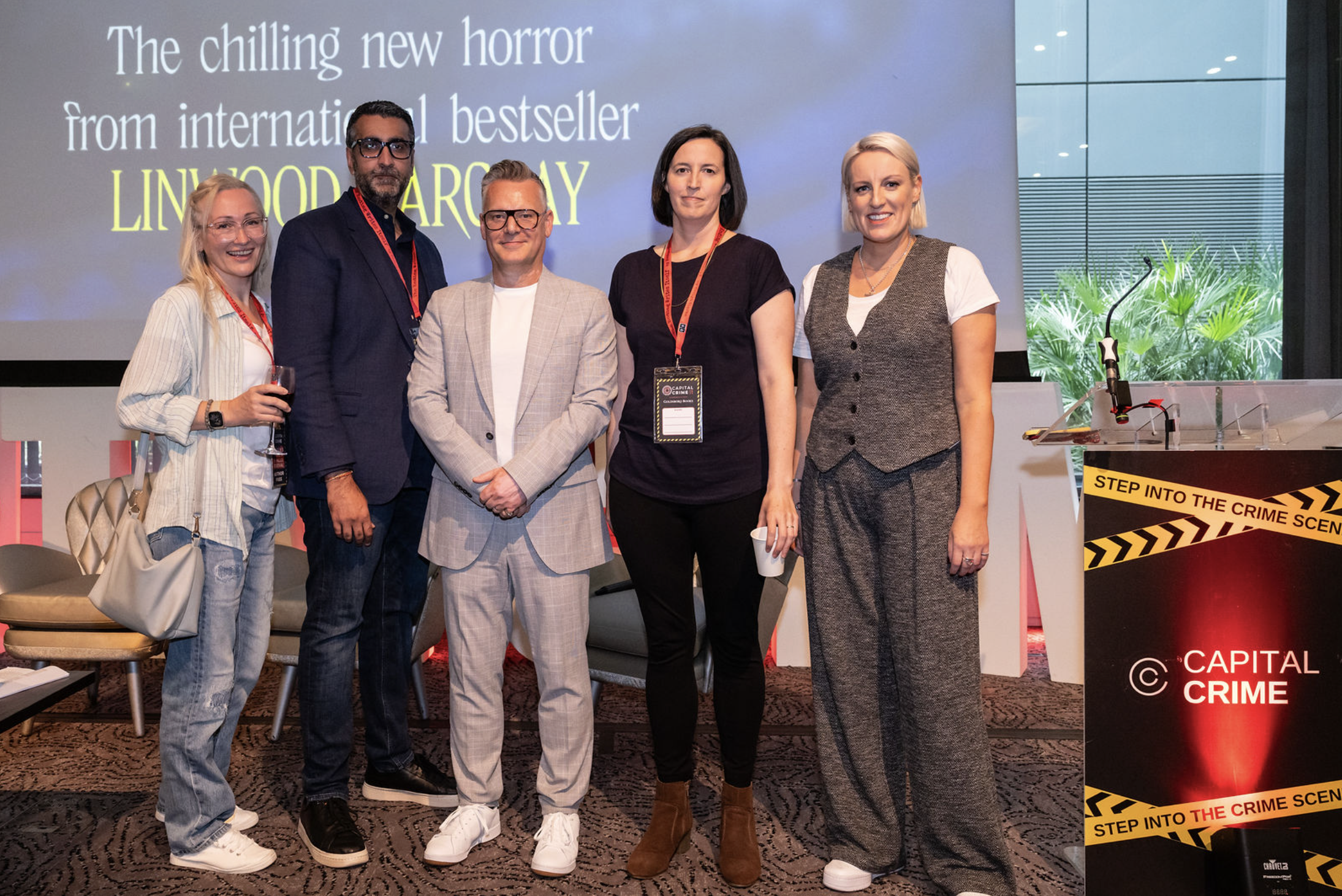Group of five people standing together on stage at a conference, with a large screen behind them displaying text about a book titled 'Lynwood Arcery.' There is a podium with crime scene tape and a sign in the foreground that says 'Step Into the Crime Scene' and 'Capital Crime.' The group includes three women and two men, all dressed in business casual or professional attire, smiling at the camera.