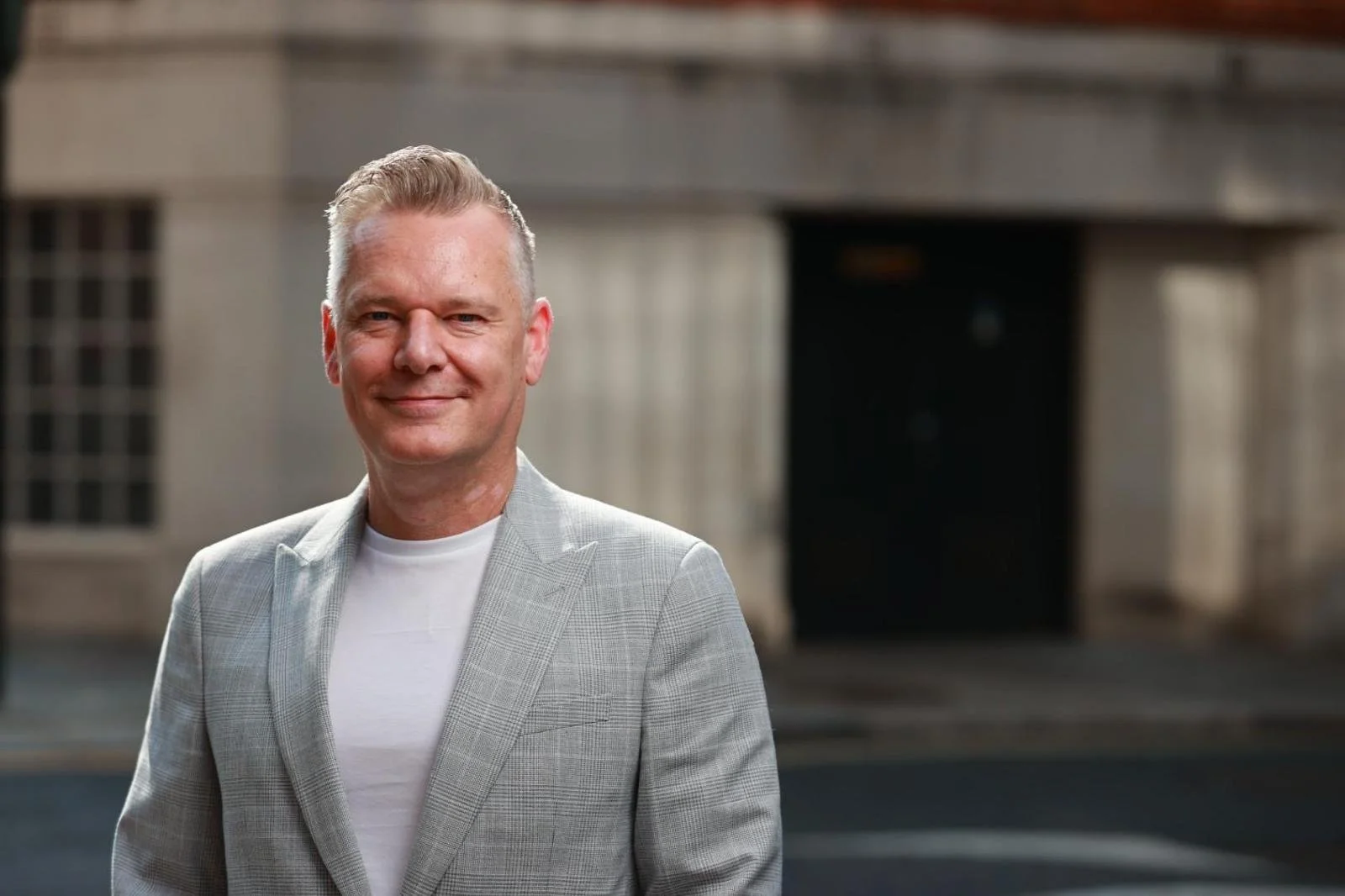 A middle-aged man with blonde hair smiling, dressed in a light plaid blazer and white t-shirt, standing in front of an urban building and street.