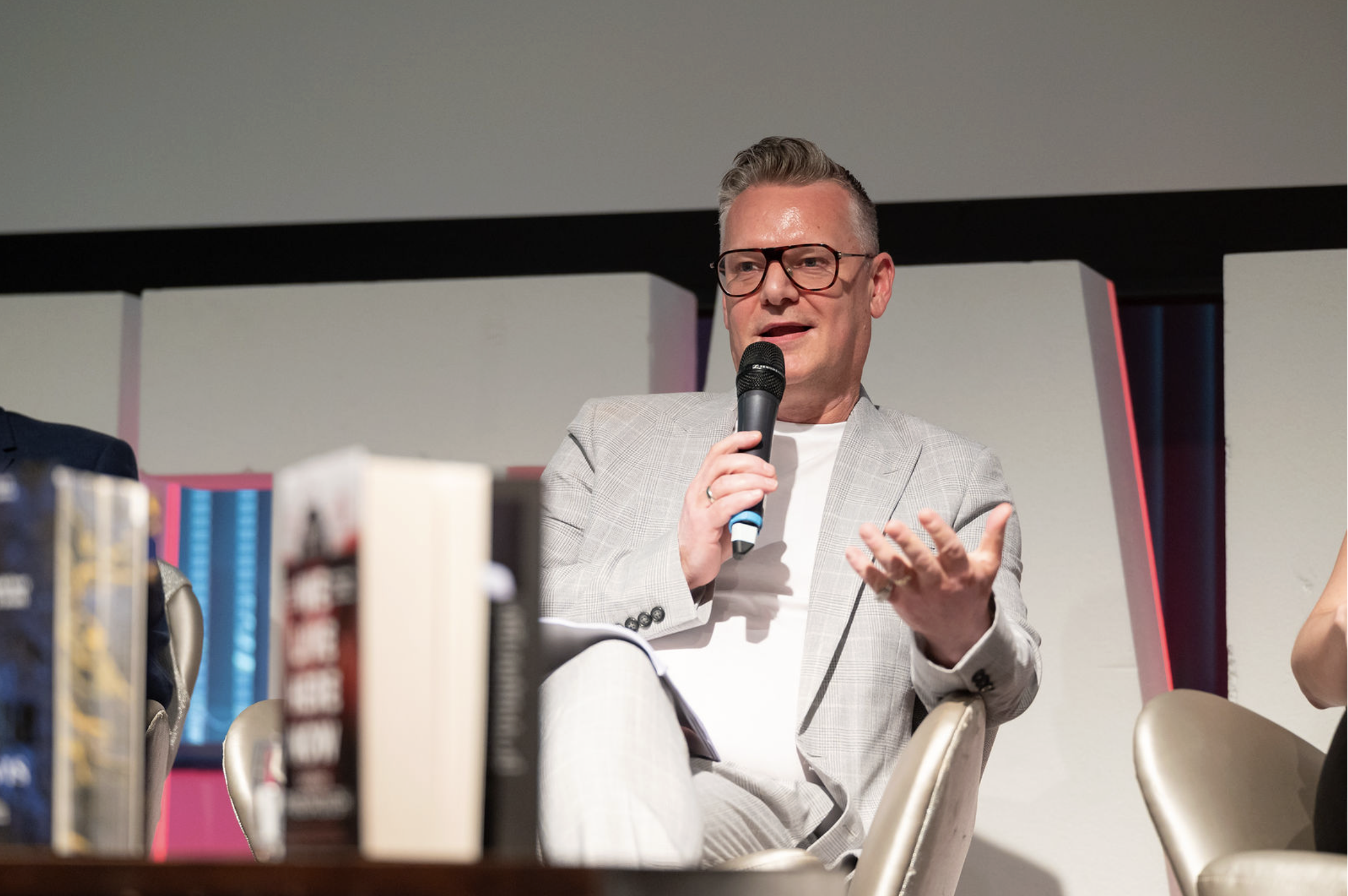 A man in a light gray suit and glasses holding a microphone and speaking at a panel discussion or conference, with books in the foreground.