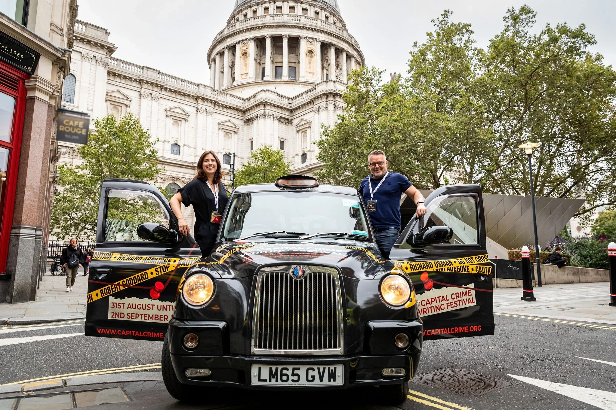 Two people standing beside a black taxi cab with crime scene tape and writing festival banners, in front of St. Paul's Cathedral in London.