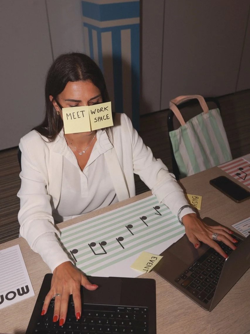Une femme assise à une table, portant un post-it sur le visage avec l'écriture "MEET WORK SPACE". Elle utilise un ordinateur portable et une tablette, avec des étiquettes et des papiers autour d'elle indiquant un environnement de travail ou d'événement.
