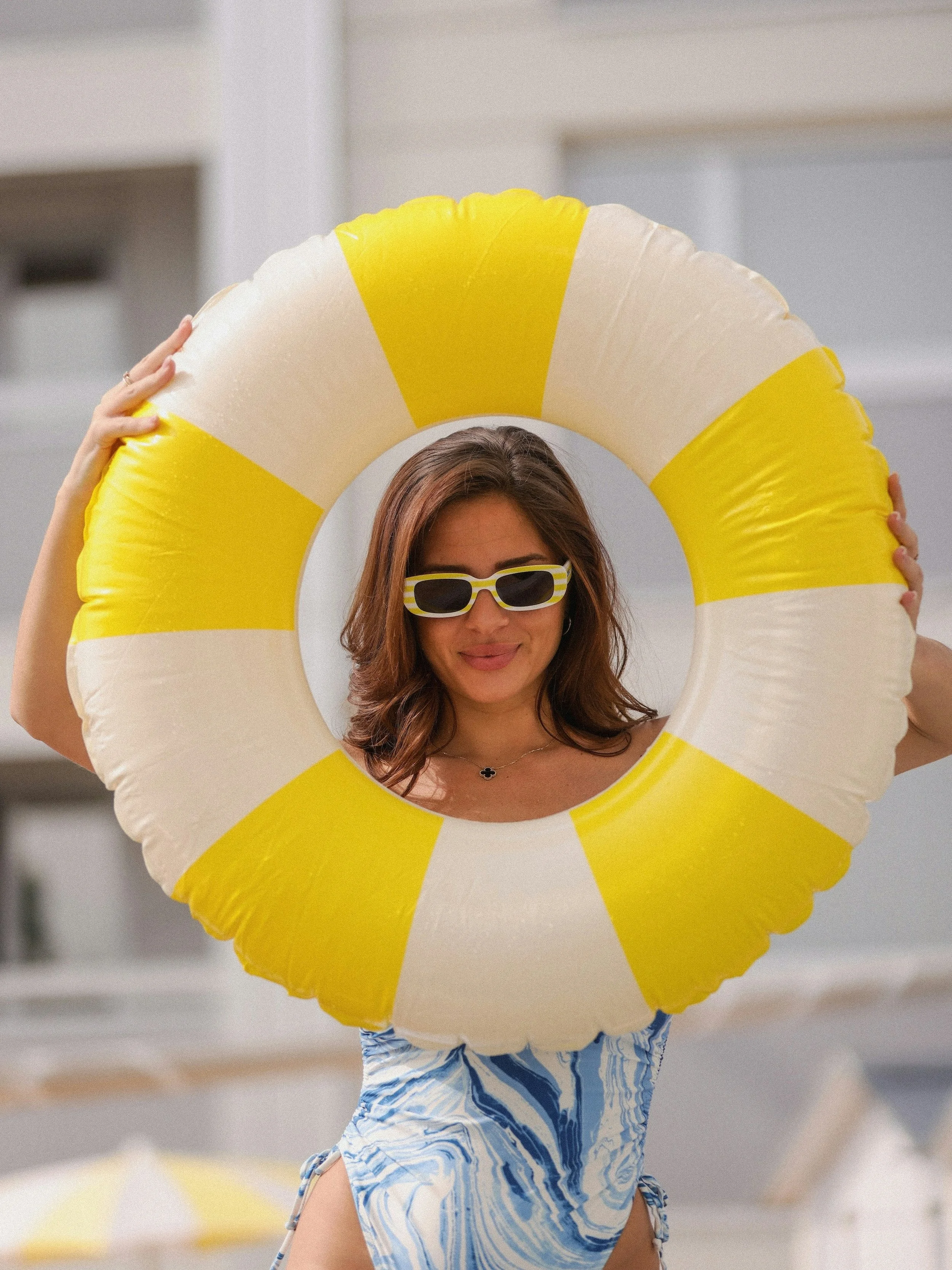Une femme portant des lunettes de soleil et un maillot de bain à motif bleu, tenant une bouée en forme de tranche de citron jaune et blanc, sur la plage.