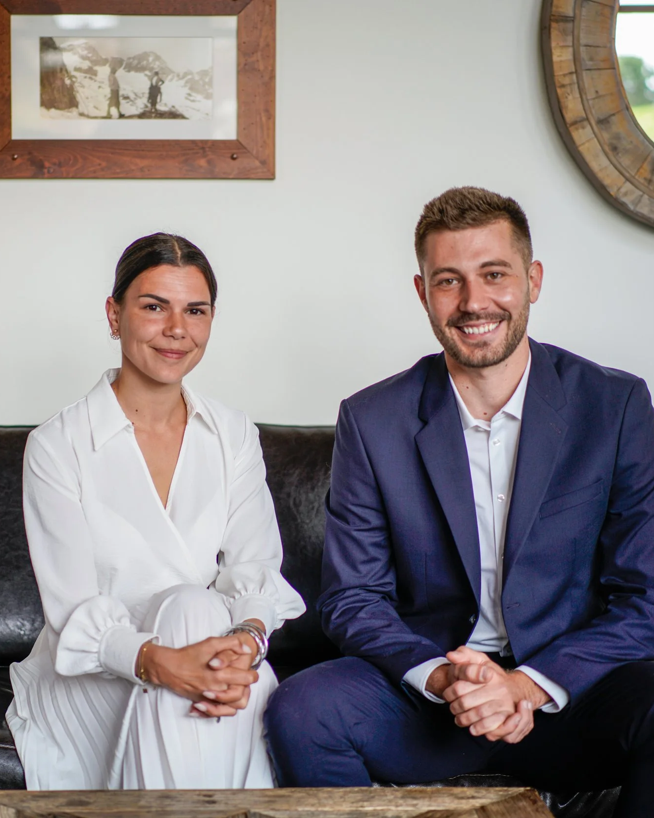 Deux personnes assises sur un canapé en cuir noir dans une pièce, une femme en blouse blanche et un homme en costume bleu, tous deux souriants.