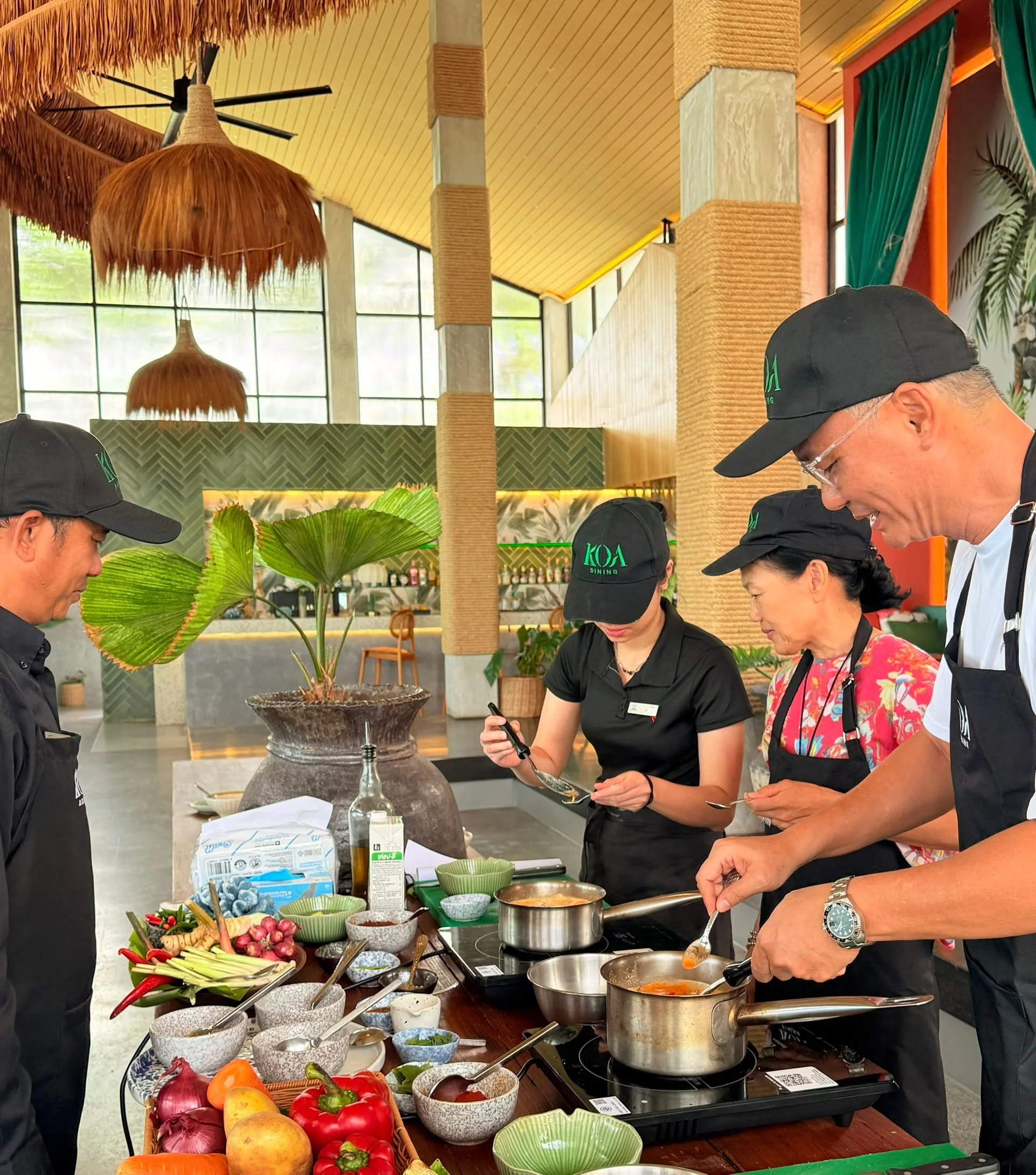 Four people cooking and preparing food in a modern, tropical restaurant setting with large windows, greenery, and hanging lamps.