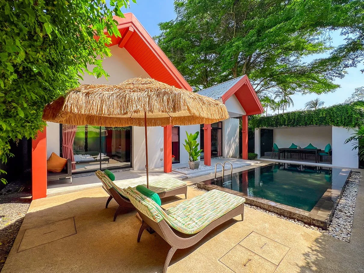 A backyard scene featuring a swimming pool with a sunbed and a straw umbrella, surrounded by green trees and a modern house with sliding glass doors.