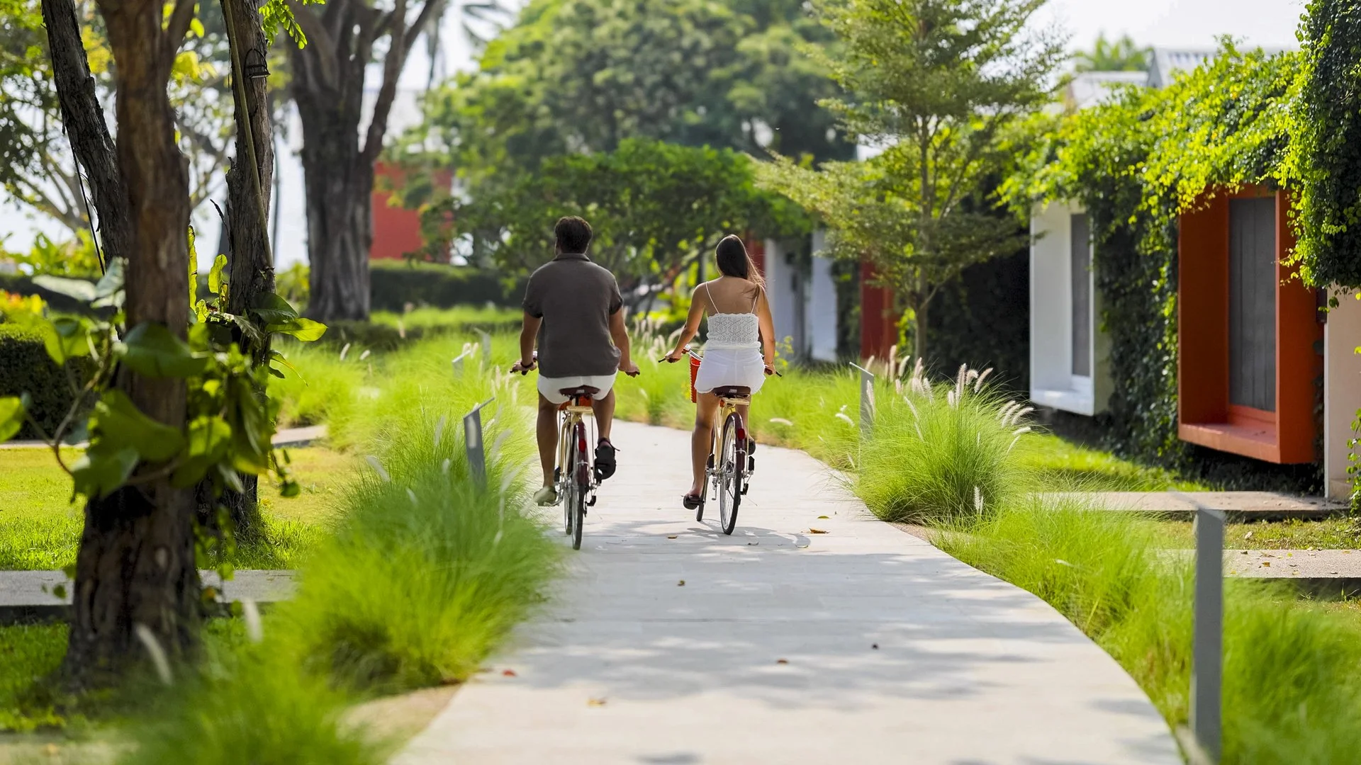 A man and woman riding bicycles on a paved path through a lush, green park with trees and plants, sunny weather.