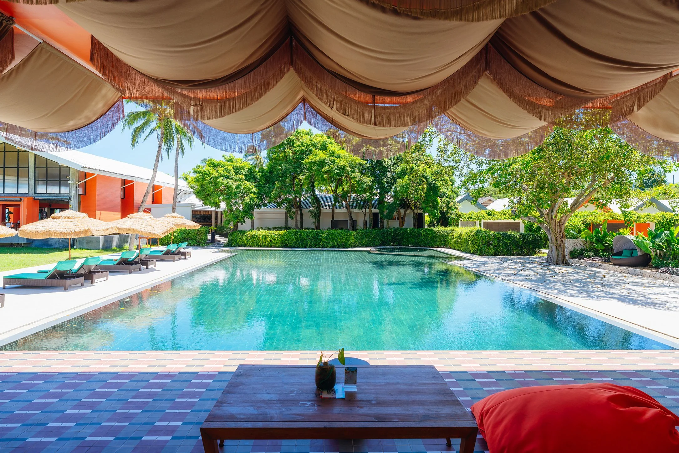 View of a swimming pool in a tropical resort, with lounge chairs and umbrellas on the side, surrounded by lush green trees and a building in the background, viewed from under a large draped canopy.